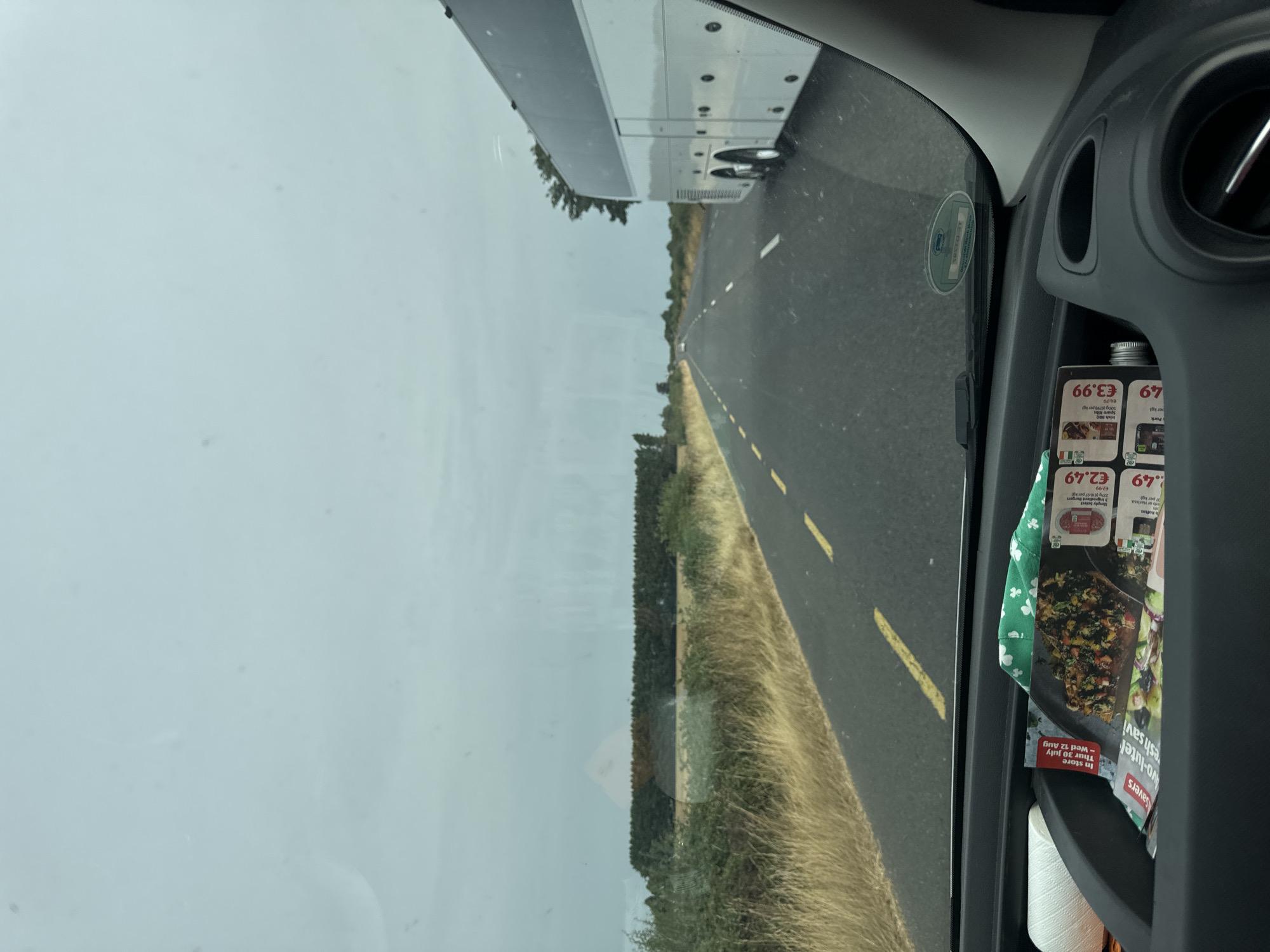 The image, taken from inside a vehicle, captures a view of a two-lane road in Grovine, Ireland. The road is bordered by dry, yellowed grass and scattered green bushes on the left side, with a dense line of trees in the distance. On the right side of the road, a white tour bus is traveling in the same direction as the vehicle from which the photo is taken. The sky is overcast with a pale blue-grey hue.

In the foreground, inside the vehicle, the dashboard is visible. A black storage compartment contains several items: a roll of white thread on the left, a green cloth with a shamrock pattern, and a stack of folded brochures or flyers. One flyer prominently displays an image of food and price tags with the currency symbol "€".