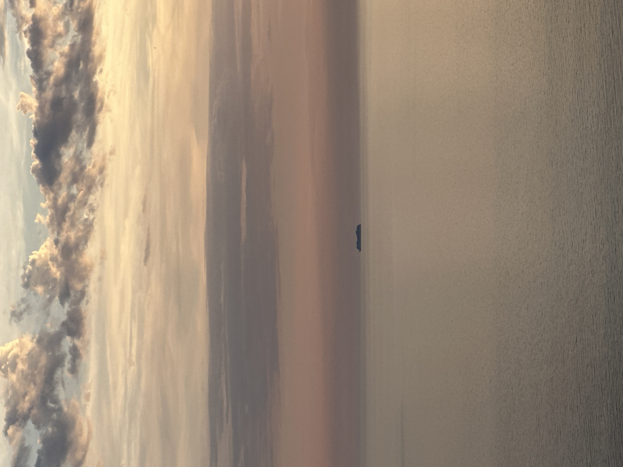The image captures a tranquil seascape at either sunrise or sunset over the Adriatic Sea, observed from Rijeka Reževići, Montenegro. The sky, occupying the upper two-thirds of the frame, displays a gradient of soft colors transitioning from light yellow and muted blue at the top, through soft orange and pink, to a hazy purplish-grey just above the horizon. Patches of cumulus clouds are visible, their edges illuminated by the low sun, creating a subtle contrast against the softer sky. Centered horizontally on the horizon line is the distant silhouette of a large vessel, consistent with a ship or ferry, appearing as a dark, indistinct form against the atmospheric haze. The sea surface, comprising the lower third of the image, is calm with subtle ripples, reflecting the subdued light from the sky in muted grey and brown tones. No individuals, landmasses, or other specific objects are present in the foreground or midground. The scene conveys a sense of serene vastness.