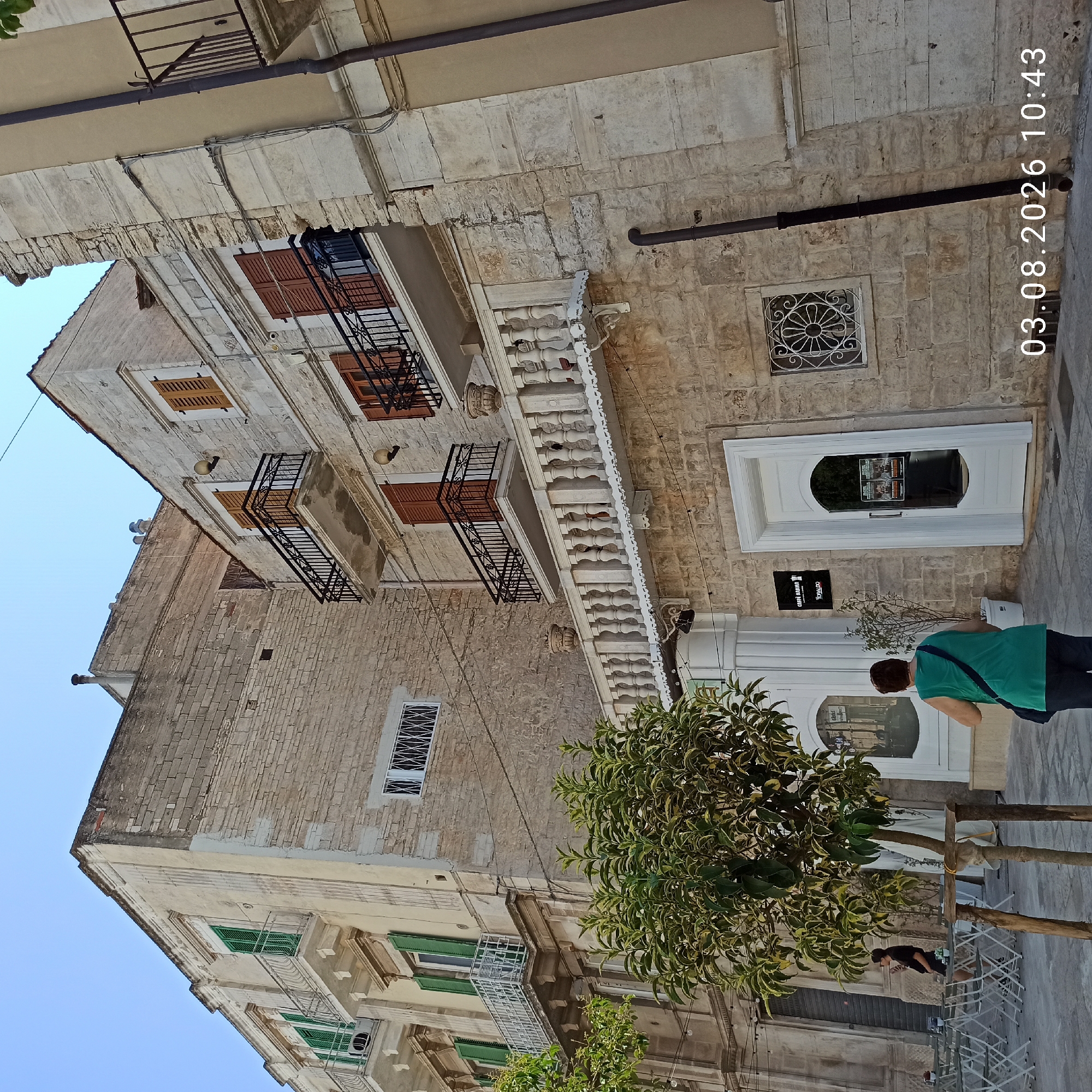 The image depicts the exterior of a building with stone and painted facades, located in the city of Ruvo di Puglia, Italy. The building features multiple stories with balconies, ornate railings, and wooden shutters on some windows. A prominent white-railed balcony with decorative urns runs along the middle level of one section of the building. The ground floor has a white door with a arched glass panel, and to its left, another arched window with a reflection. A sign for "CAFFÈ ANIMA" is visible above the door.
In the foreground, a young person wearing a black t-shirt and shorts is seated at an outdoor table, looking at their phone. A woman in a green sleeveless top and dark pants is standing and facing away from the camera, holding a dark bag over her shoulder. A small, leafy tree stands in a wooden frame near the entrance. The ground is paved with stone tiles. A date and time stamp "03.08.2026 10:43" is visible in the lower right corner of the image. The sky is clear and blue.