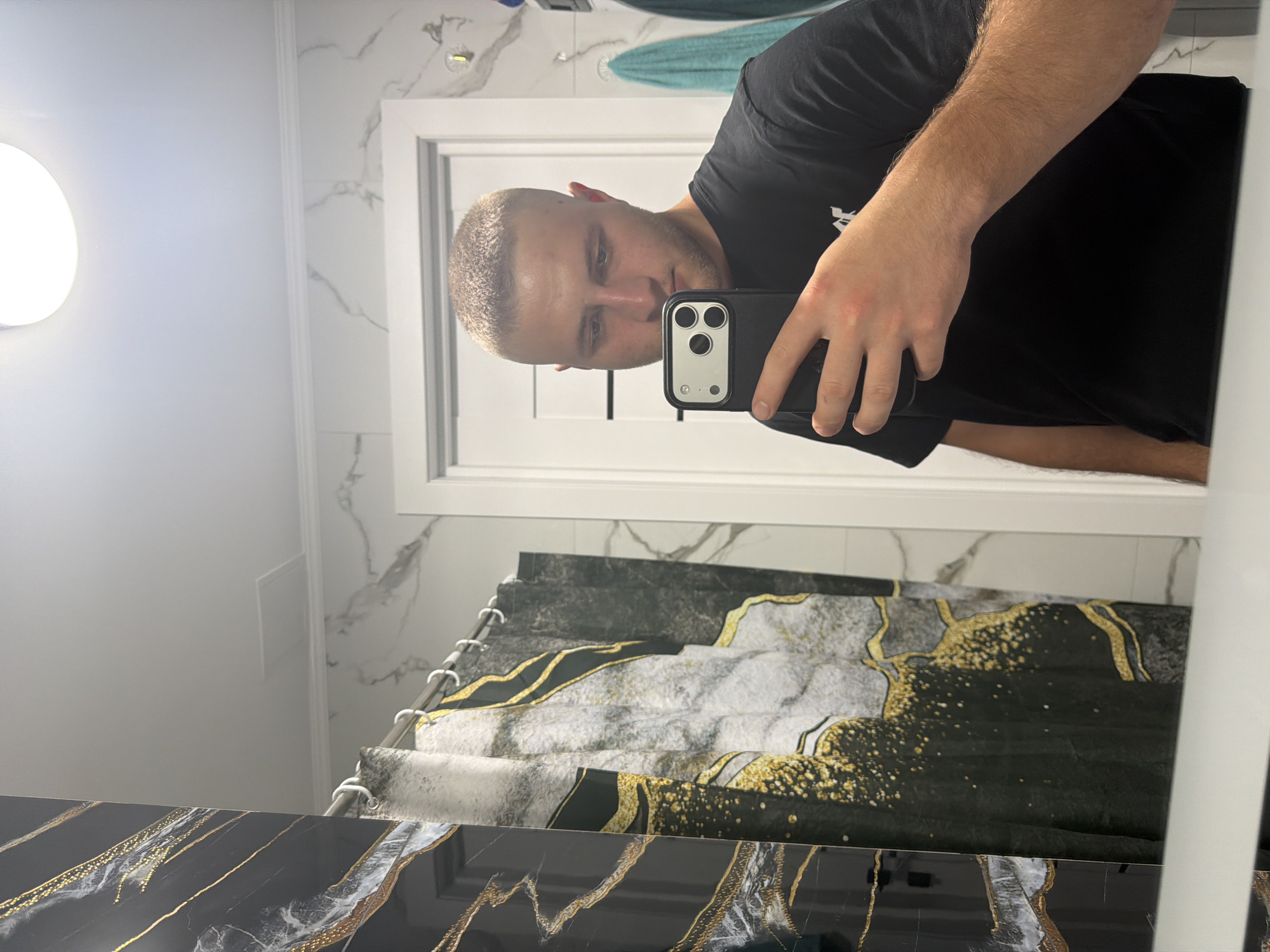 A young Caucasian male with a buzz cut and a black t-shirt is taking a mirror selfie in what appears to be a bathroom. He is holding a black smartphone in his right hand, angled to capture his reflection. The background reveals a modern bathroom with marble-patterned walls, a white door, and a shower curtain with a black, gray, and gold abstract design. Two teal towels are visible hanging on the wall to the right. The lighting in the room comes from a circular ceiling light. The scene is devoid of any specific identifying markers that would place it in Strășeni, Moldova.