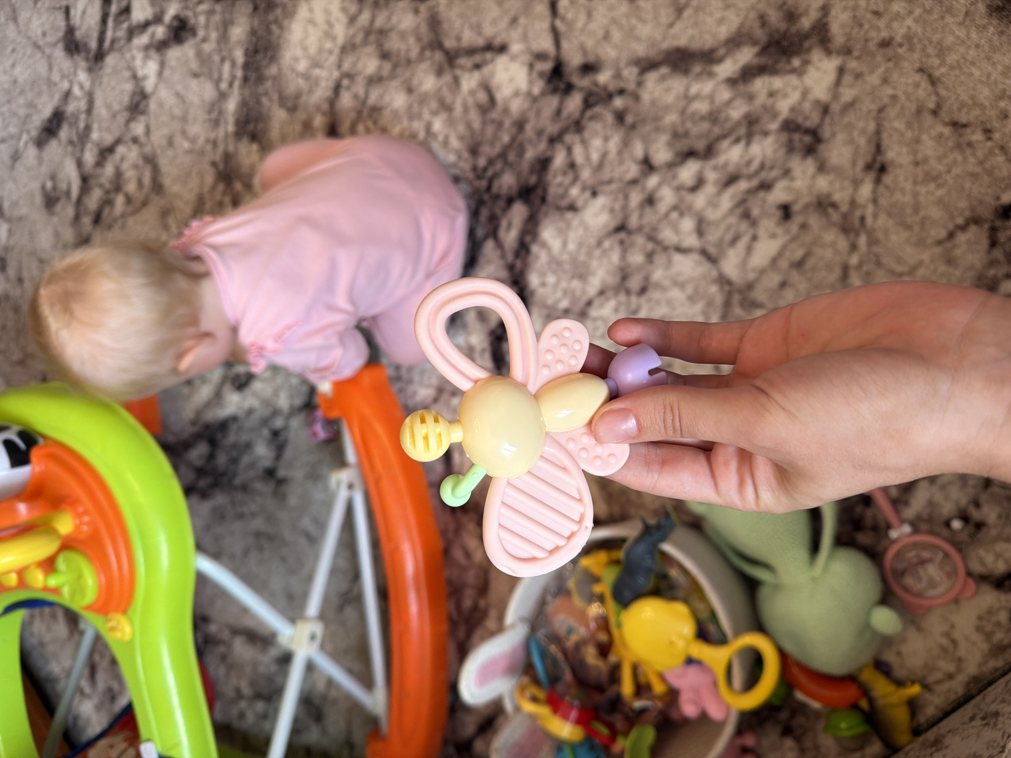 In Sovkhoza "Razdol'e", Russia, a person's hand is holding a pastel pink and yellow butterfly-shaped baby rattle. The rattle has textured wings and a circular handle. In the background, a baby wearing a pink outfit is seated on a textured rug, facing away from the camera, and appears to be playing with a green toy. A large, multi-colored baby activity center is partially visible to the left of the frame, and a white bowl filled with various baby toys is also present. The environment appears to be an indoor setting, likely a home.