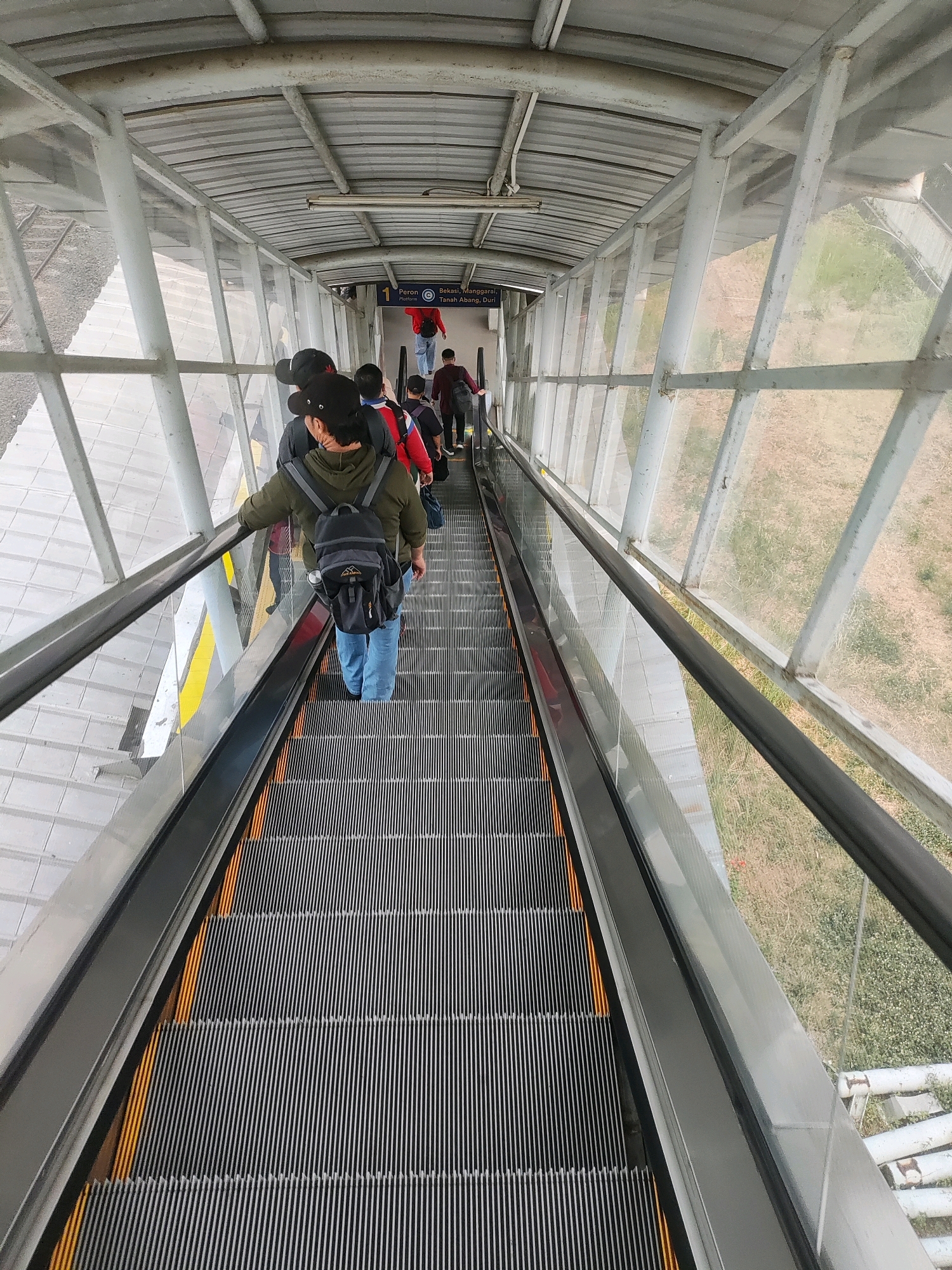 The image captures an overhead perspective of a covered escalator at a train station in Telagamurni, Indonesia. Several individuals are ascending the escalator, some carrying backpacks. The escalator is enclosed by a metal and glass structure, with an exterior view of an outdoor area with vegetation. A sign indicating "Peron 1 Platform" and destinations "Bekasi, Manggara" suggests this is a functional transportation hub. The lighting appears to be a mix of natural light filtering through the glass and artificial overhead lighting. The scene is orderly, with people moving in a single direction.