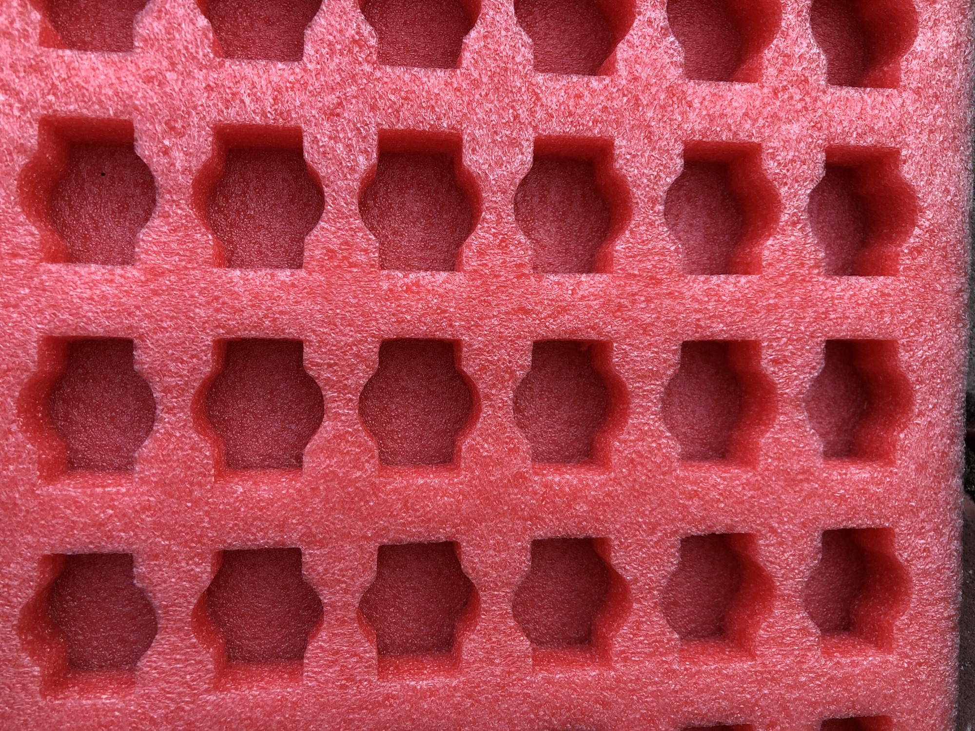 The image shows a close-up, overhead view of a textured red foam insert. The foam has a grid of octagonal indentations, arranged in rows and columns. The indentations are deep enough to create shadows within them, giving the impression of multiple dark pockets within the red foam. The texture of the foam is slightly rough and porous. The lighting appears to be from above and slightly to the side, casting subtle shadows. The scene is static and shows no signs of activity. There are no people or animals present. The location in the city of Московский Ветеран Снт, Russia, is not visually discernible from the image.