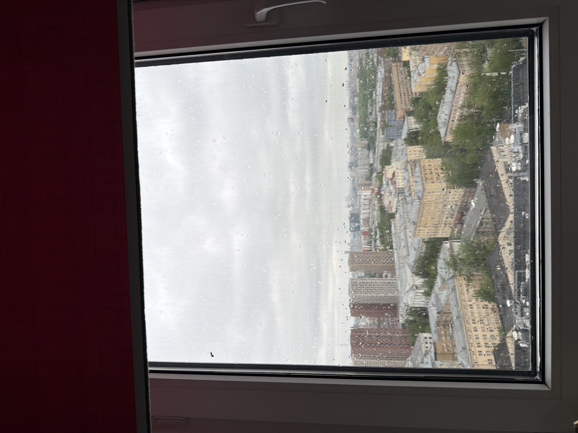 The image shows a view from a window in Unknown, Unknown on a rainy day. Raindrops cover the windowpane, partially obscuring the urban landscape outside. The buildings are a mix of residential and commercial structures, with varying heights and architectural styles. Some greenery is visible, suggesting trees or parks interspersed within the city. The sky is overcast and grey, indicating a rainy and gloomy weather condition. To the left of the window, there's a partial view of a red wall.