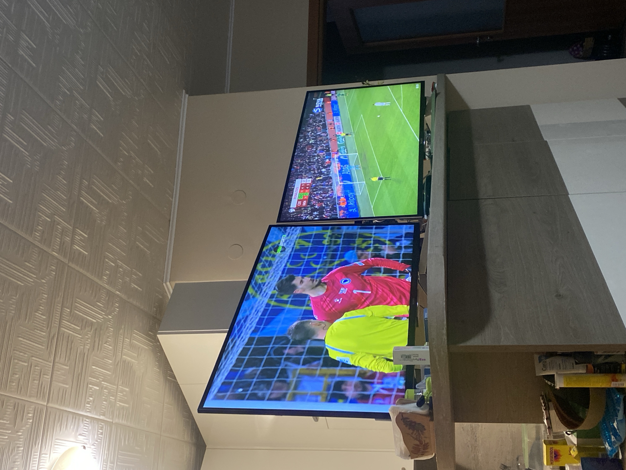 The image shows two televisions stacked on top of each other in what appears to be an indoor environment. Both screens are displaying a soccer match; the upper screen shows a wide view of the field during gameplay, while the lower screen features a close-up view of a goalkeeper in a pink jersey.
It is not possible to determine from the image whether the location is in Pleven, Bulgaria.