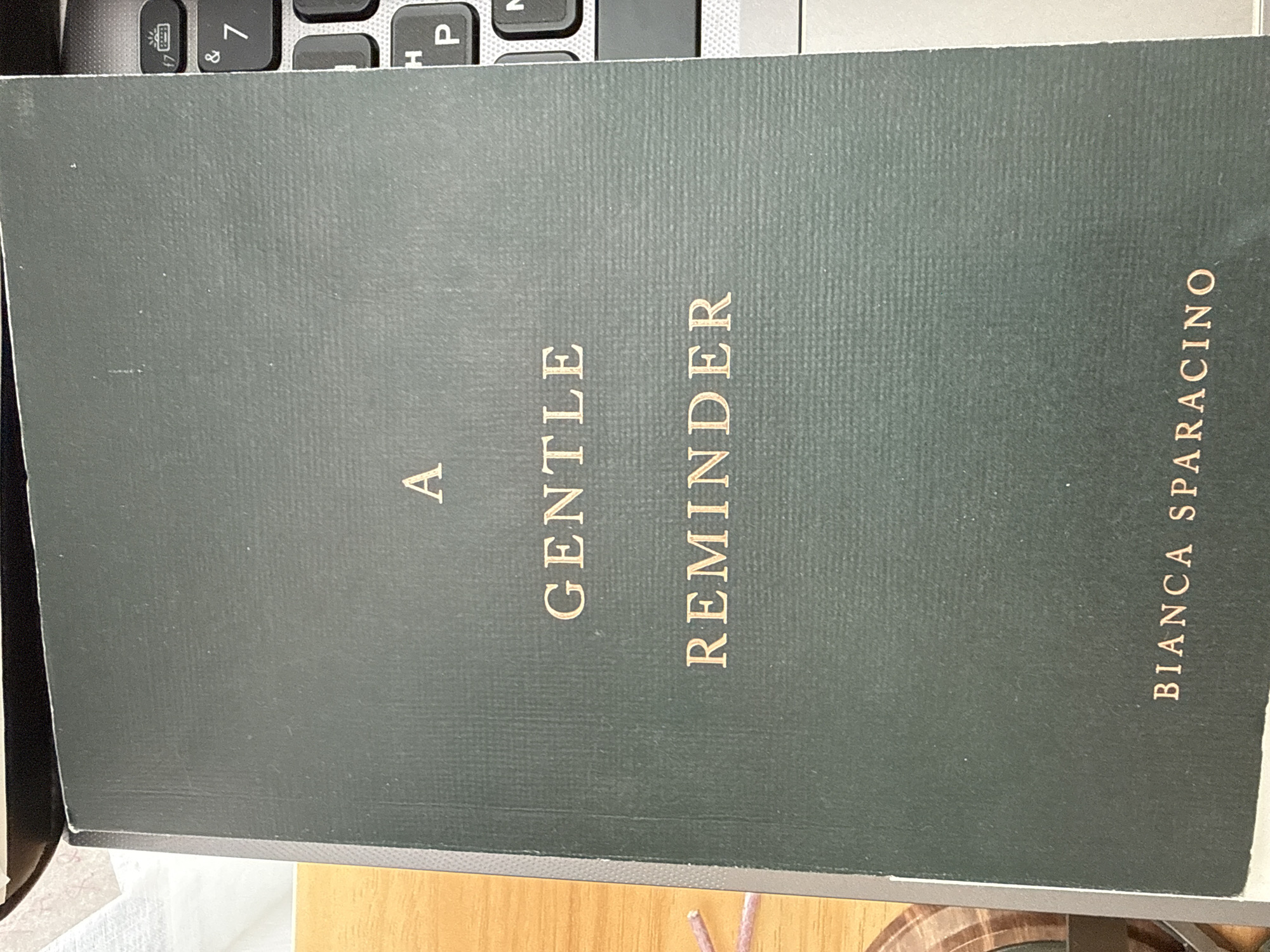 A book with a dark green cover is lying on a surface, possibly a desk or table, with a laptop visible to the right of the book. The book's title, "A GENTLE REMINDER," is printed in gold sans-serif font. Below the title, the author's name, "BIANCA SPARACINO," is also printed in gold. The book appears to be a paperback. The background includes a portion of a wooden surface to the left and a keyboard of a laptop to the right. The image is taken from a slightly overhead angle. There is no discernible information to ascertain the specific location in Stock Island, United States of America.