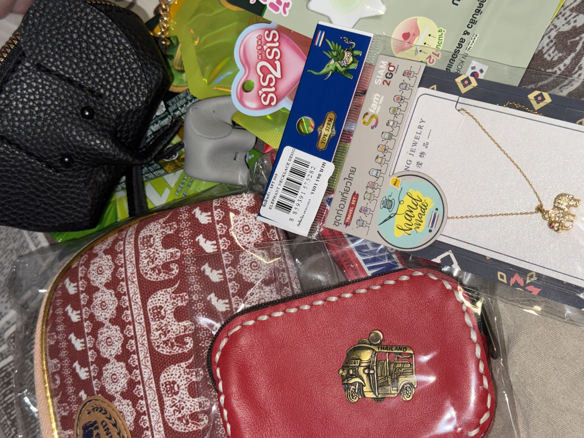 The image shows a collection of Thai souvenirs. Items include a black leather purse, a red and white elephant-patterned coin purse, a small red coin purse with a gold Tuk-Tuk emblem, elephant-themed necklace, a box of Yok Siam stamps, sis2sis lip product, and Siam 2GO travel set. The image appears to be taken indoors. The city of Perm, Russia, is not present in the image.