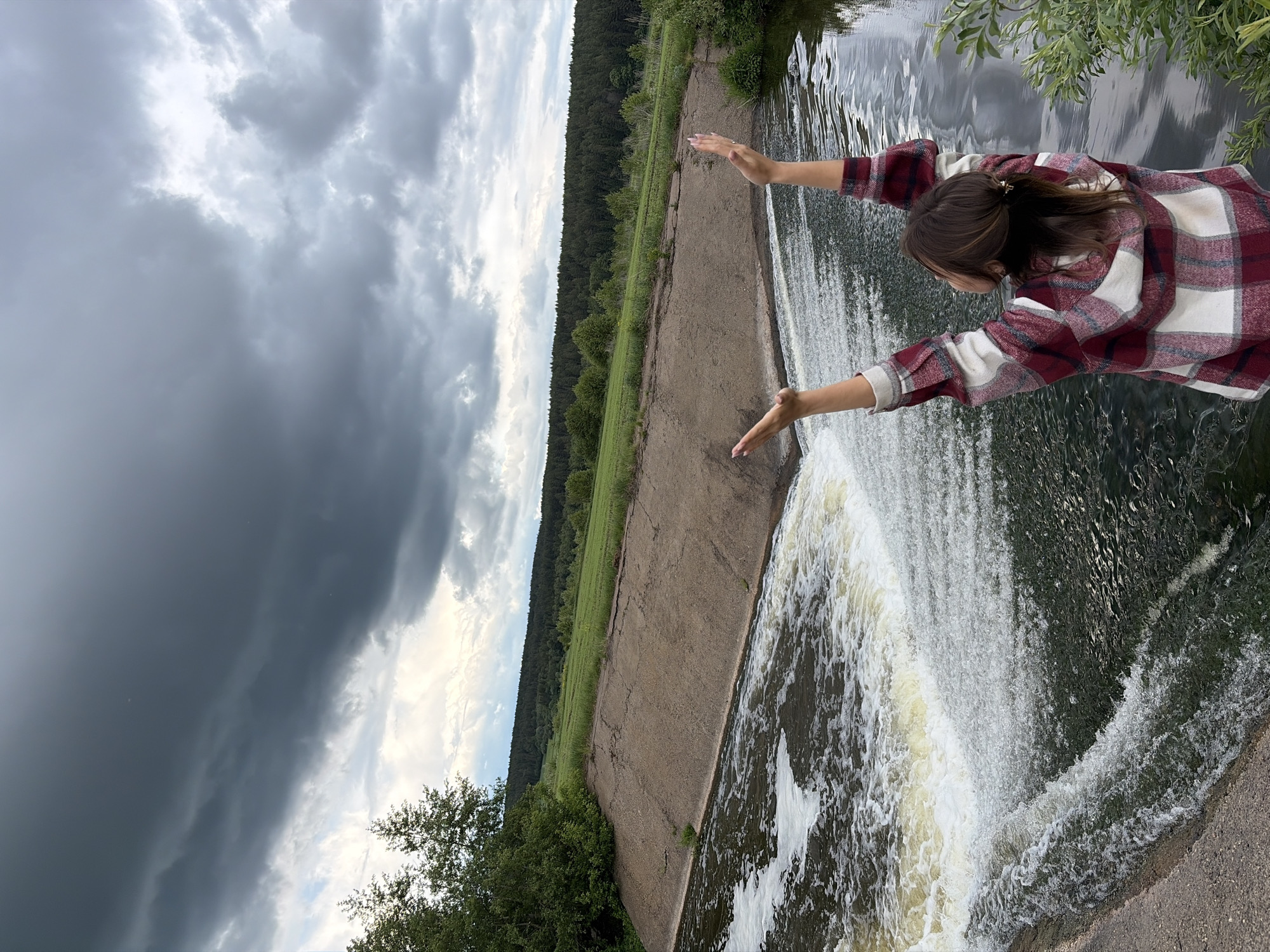 The image captures a scene at a waterfall in Nizhnii Vishur, Russia. A woman in a red and white plaid shirt stands at the edge of a concrete structure, presumably a dam, reaching her arms out towards the cascading water. The waterfall is the primary focal point. Behind the dam is a grassy field and a line of trees, suggestive of a forest. Above, a cloudy sky dominates the view. The overall perspective suggests the photo was taken at an angle.