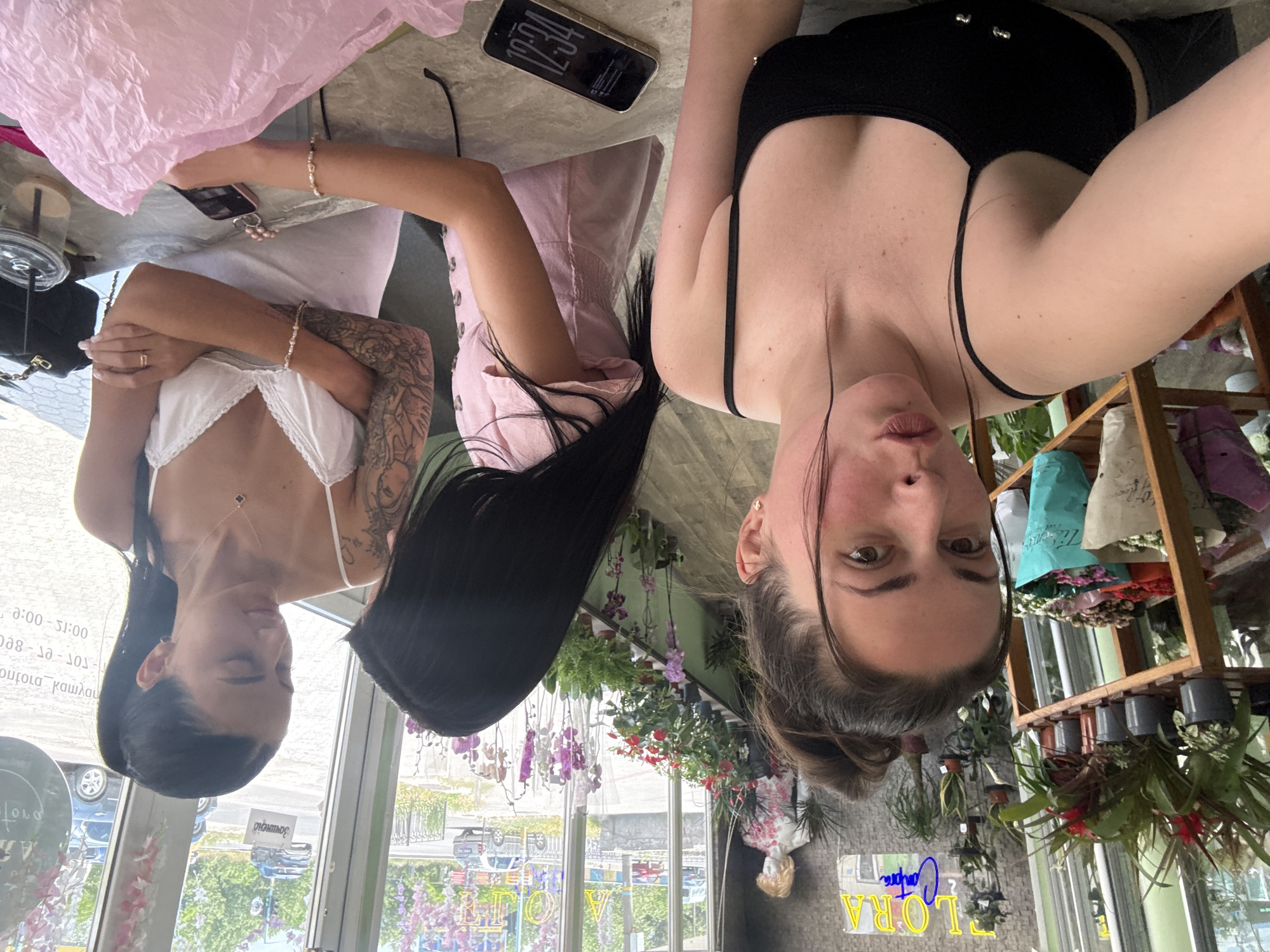 Three women are posing for a selfie in what appears to be a flower shop. The woman in the foreground is wearing a black spaghetti strap tank top and has her hair pulled up in a messy bun. She is looking directly at the camera with her mouth slightly open. To her right, another woman is wearing a light pink dress and has her back mostly to the camera. The third woman is on the right side of the image, wearing a white camisole and looking down. She has tattoos on her arm and is wearing a necklace. In the background, there are shelves filled with plants and flowers, as well as a mannequin. Through the large windows, a street scene with cars and buildings is visible. A table in front of the women has phones and other items on it.