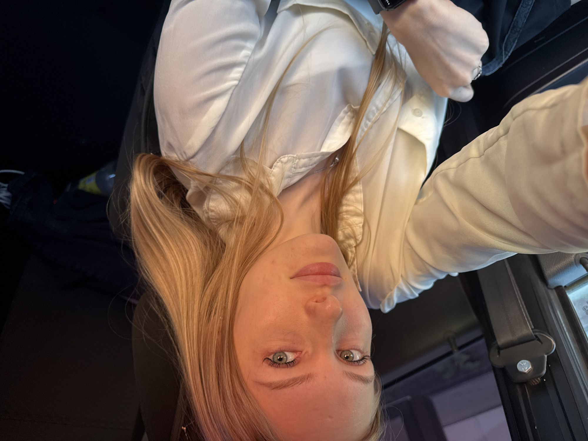A blonde woman is taking a selfie while seated in a car. She is wearing a white button-down shirt and a wristwatch. A seatbelt is visible. The location is potentially in Perm, Russia, based on the car's interior and general environment, though there are no definitive local landmarks visible.