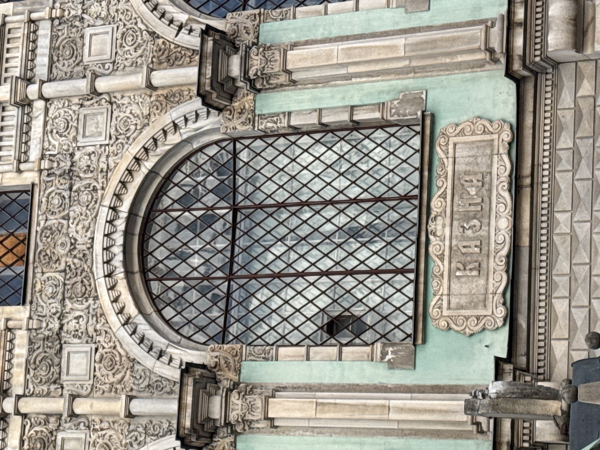 The image shows a section of the architectural facade of the Bolshoi Theatre in Moscow, Russia. The facade exhibits ornate detailing, including sculpted stone elements, decorative moldings, and a large arched window with a diamond-patterned grid. A sign with Cyrillic script is visible adjacent to the window, and the overall color palette is a combination of cream, gray, and hints of light green.