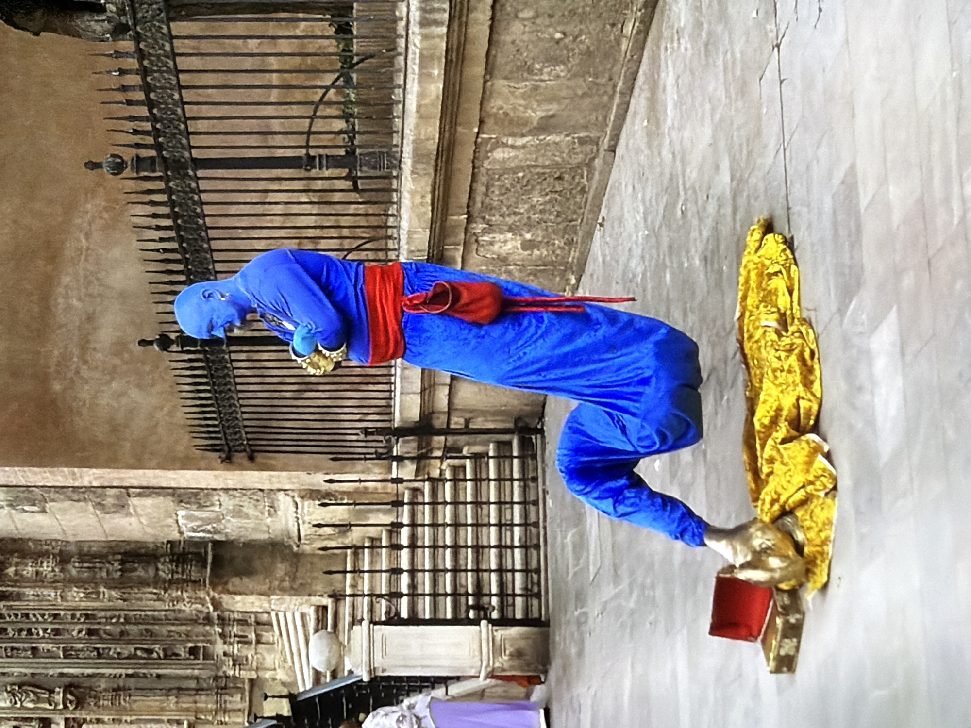 This image captures a street performer dressed in a bright blue full-body suit, complete with a red sash and a matching red pouch tied around their waist. Their face is also covered in blue, creating a striking visual. The performer is in a contorted pose, with their legs bent and twisted, appearing to levitate or defy gravity. They are holding a golden object in their right hand, which is resting on a red and gold box. Next to them, a crumpled golden fabric is draped.
The setting appears to be an indoor or covered public space, possibly a historical building or a grand hall, suggested by the stone architecture, ornate iron gates, and intricate carvings visible in the background. The floor is made of large, light-colored tiles. The lighting suggests daytime, but it's difficult to ascertain the exact time or weather conditions. The overall impression is one of a performance or an artistic display, with the performer's unusual pose and attire designed to attract attention and evoke wonder. There is no visible text in the image.