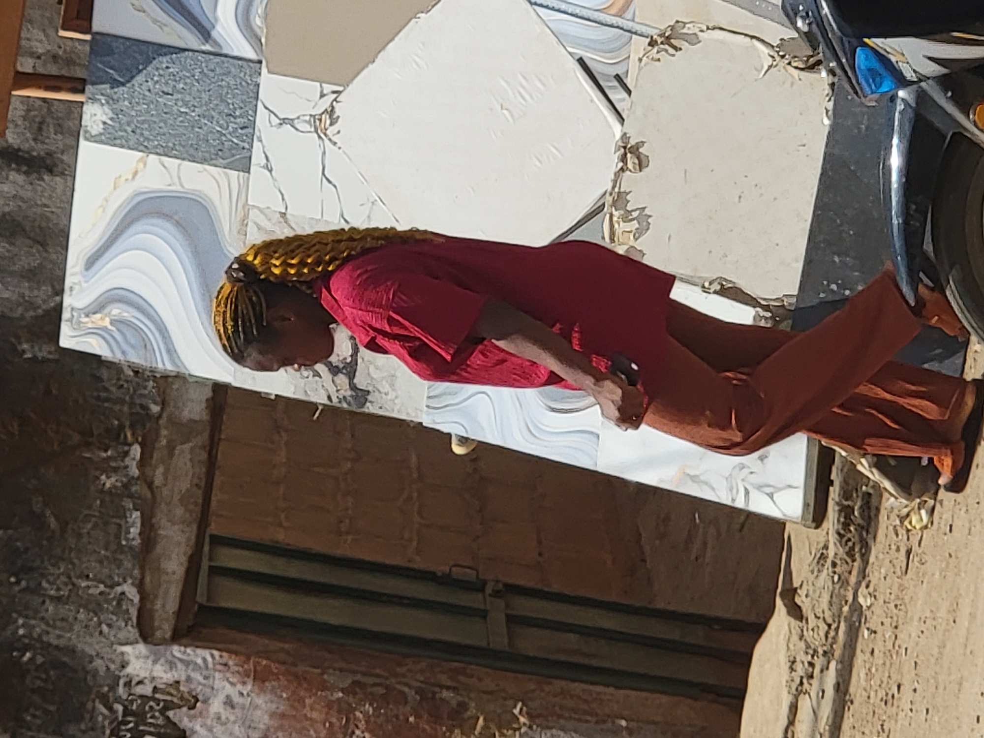 A woman with yellow braided hair stands on a variety of differently colored tiles. She wears a red shirt and orange pants. To the right of the woman is part of a motorcycle. Behind the tiles is a textured, worn wall with a dark opening. The location is Maiburiji, Nigeria.