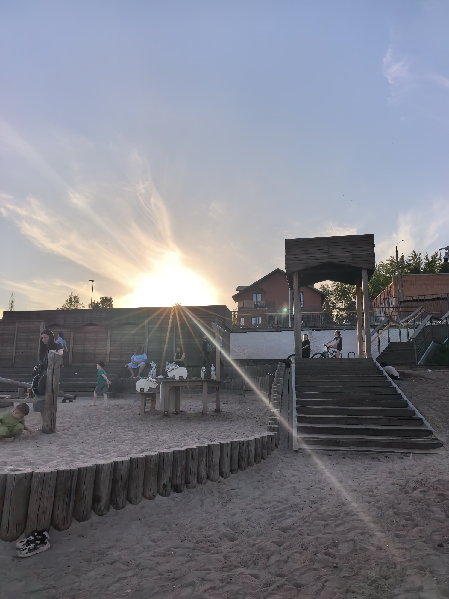 The image shows a public space in Sterlitamak, Russia, possibly a park or beach area, at sunset. The foreground is a sandy area with wooden logs forming a barrier, with children playing. Some adults are seated on a wooden structure in the background. A wooden table is visible with white objects on top. In the mid-ground, a set of wooden stairs leads up to a platform with a wooden structure on top, where a person is riding a bicycle while another stands nearby. Behind the platform is a two-story brick building. The sky is bright with the sun setting, causing a lens flare.