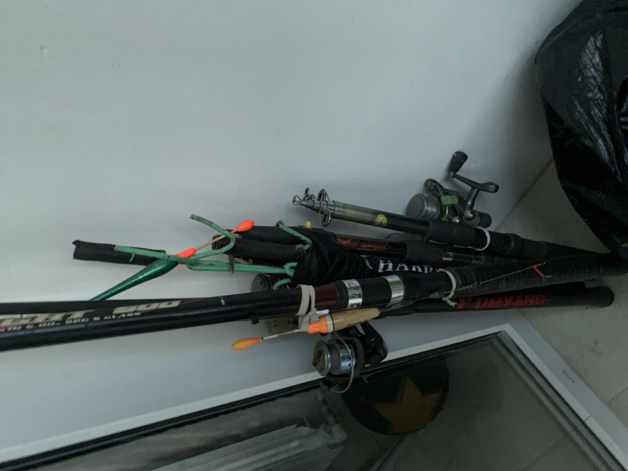 The image shows a collection of fishing rods and reels leaning against a white wall. There are at least three fishing rods visible, with one larger rod in the foreground that has a reel attached. The rods have various attachments, including orange floats and green guides. A black trash bag is visible in the lower right corner of the image. The scene appears to be indoors, possibly in a storage area or garage. The image does not provide any information about the city of Stavropol, Russia.