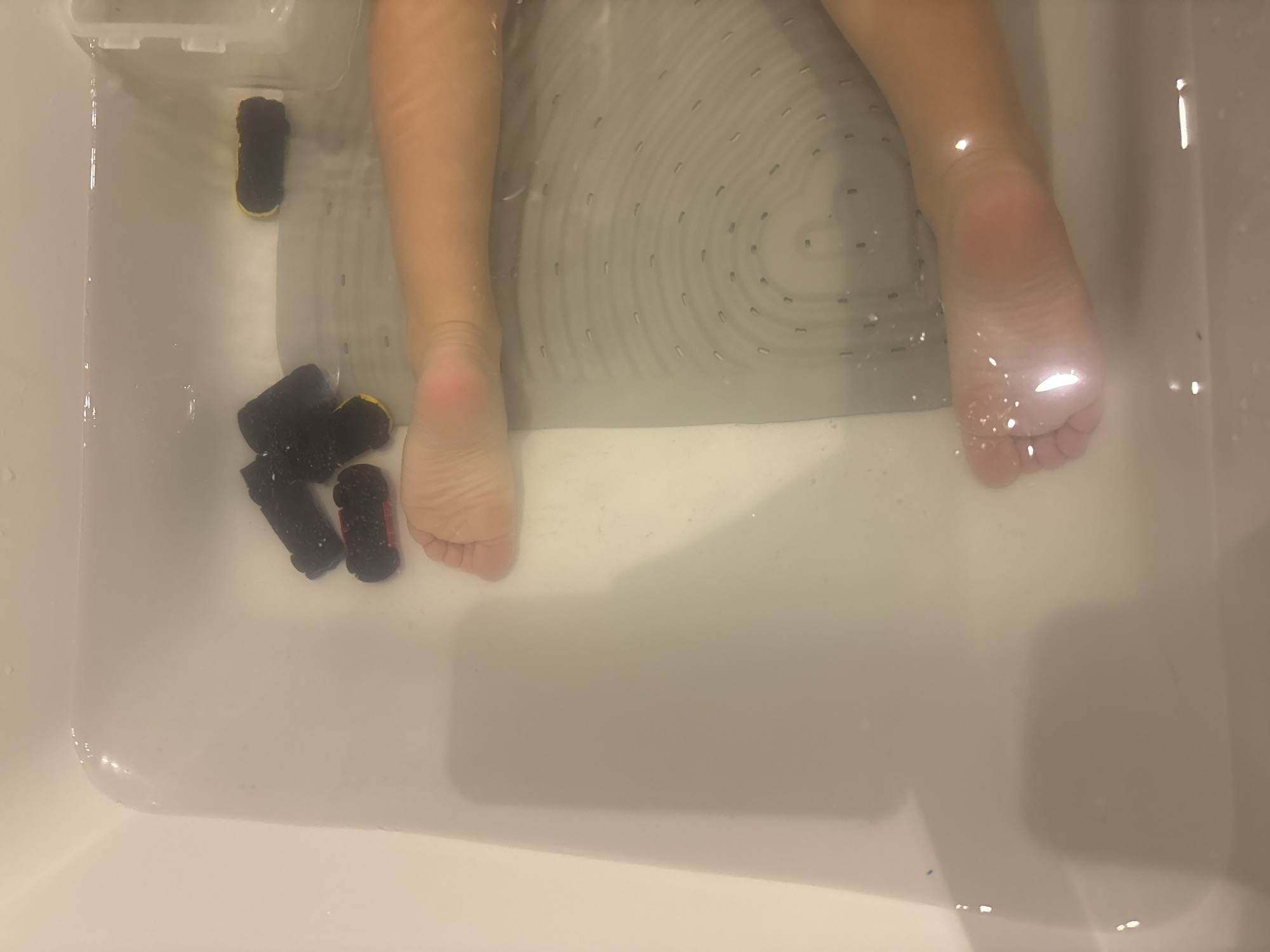 A high-angle medium shot shows a child's feet submerged in a bathtub filled with clear water. The bathtub is white ceramic with a textured, light gray mat at the bottom. Several small, black toy cars with yellow accents are floating in the water near the child's upper leg. The child's skin is peachy-toned. The lighting is artificial and appears to be overhead. The scene is an indoor bathroom setting. There is no visible indication of the location within the city of Dubai, United Arab Emirates.
