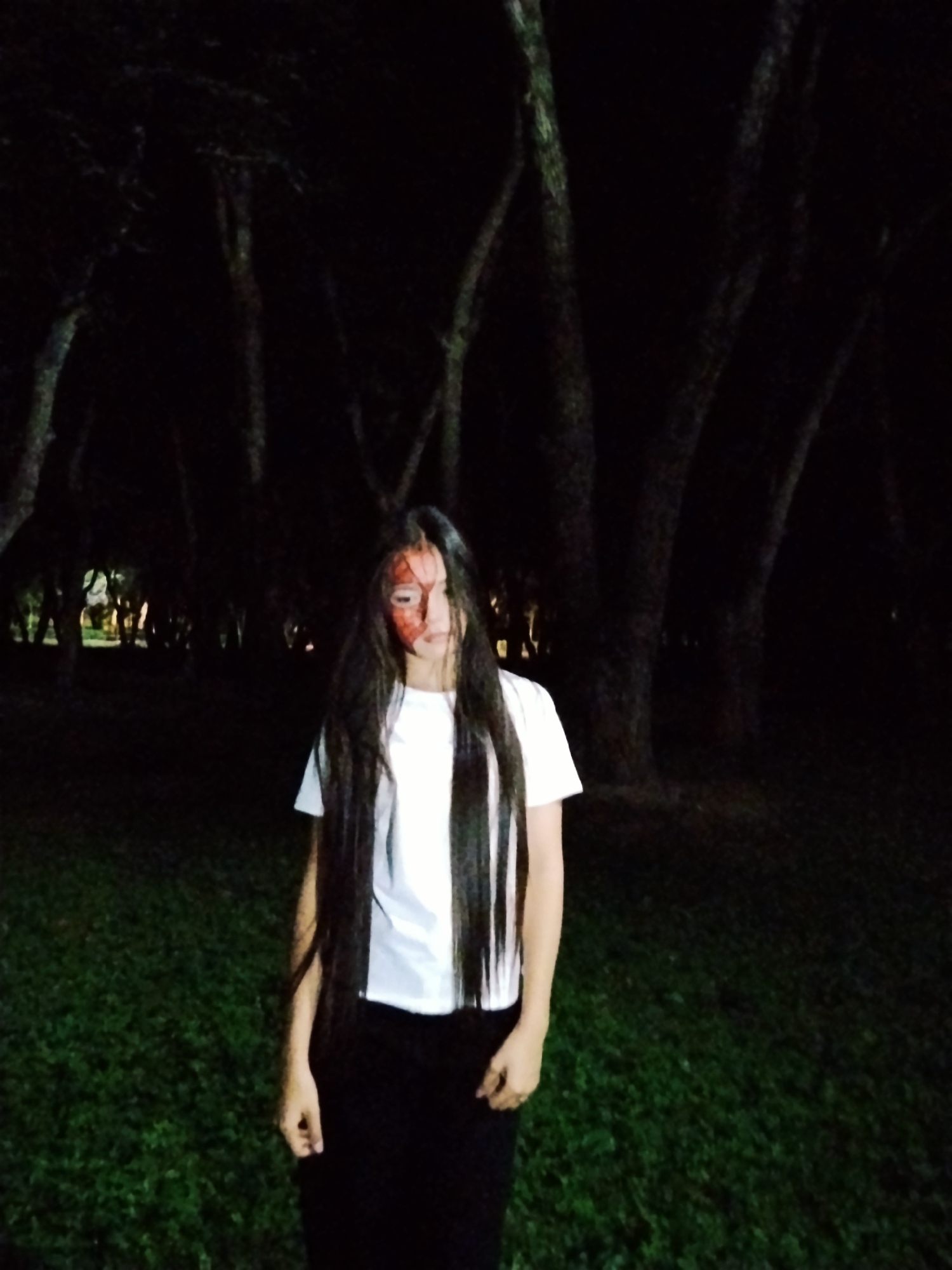 A young woman with long dark hair and what appears to be face paint, possibly simulating a bloody injury on the left side of her face, stands in a dimly lit outdoor area at night. She is wearing a white t-shirt and dark pants. The background consists of trees and grass, suggesting a park or wooded area. The lighting is low, with some light sources visible in the distance, possibly from buildings or streetlights. The scene has a dark and slightly eerie atmosphere. The location is described as being in the city of Bishkek, Kyrgyzstan.