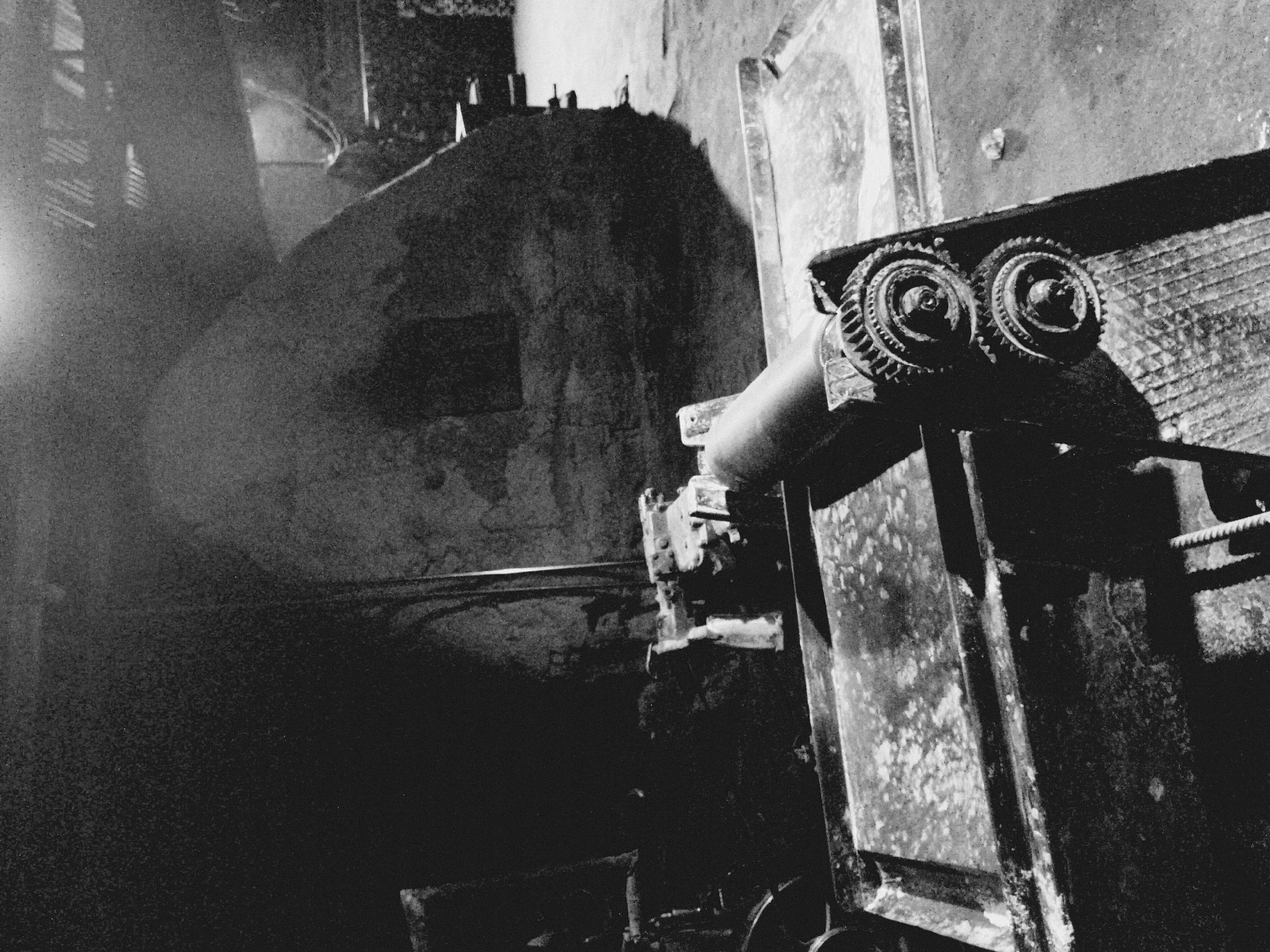 The image depicts a dimly lit, possibly industrial setting. In the foreground, a metallic structure with prominent cogwheels and a grated surface is visible. The metal appears weathered and mottled. In the background, a large, shadowed structure or wall with some markings or signs upon it. The overall ambiance is dark and conveys a sense of age and disuse. The location in the city of Unknown, Unknown is also Unknown.