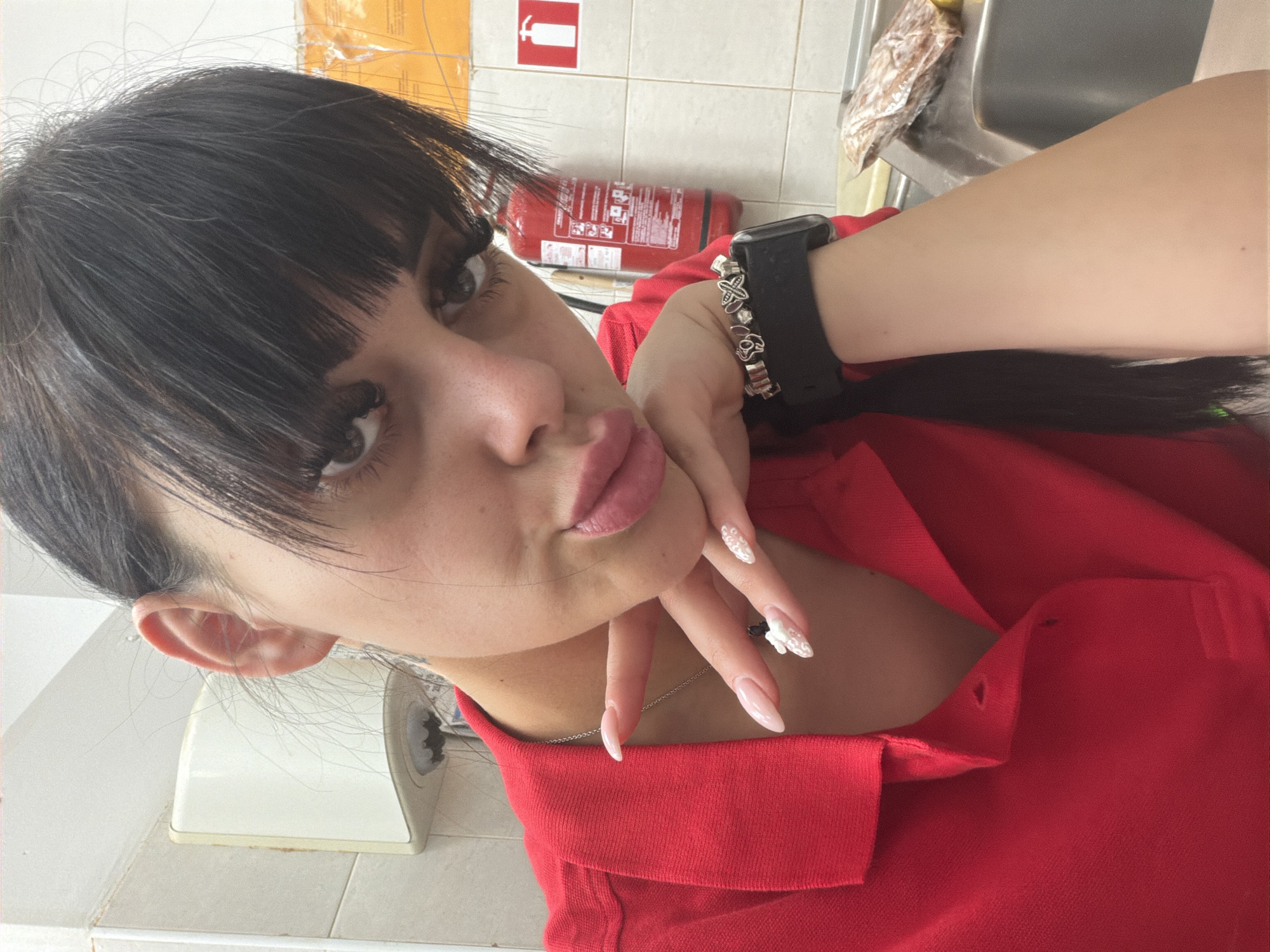 A young woman with dark hair and bangs, wearing a red polo shirt, poses for a selfie in what appears to be a commercial kitchen or utility area. She has long fingernails with white nail art. A black smartwatch is visible on her left wrist, adorned with silver charms. In the background, a fire extinguisher is mounted on a tiled wall. A sink and a packaged item are also visible. The lighting suggests an indoor setting, possibly in Plovdiv, Bulgaria, given the context.