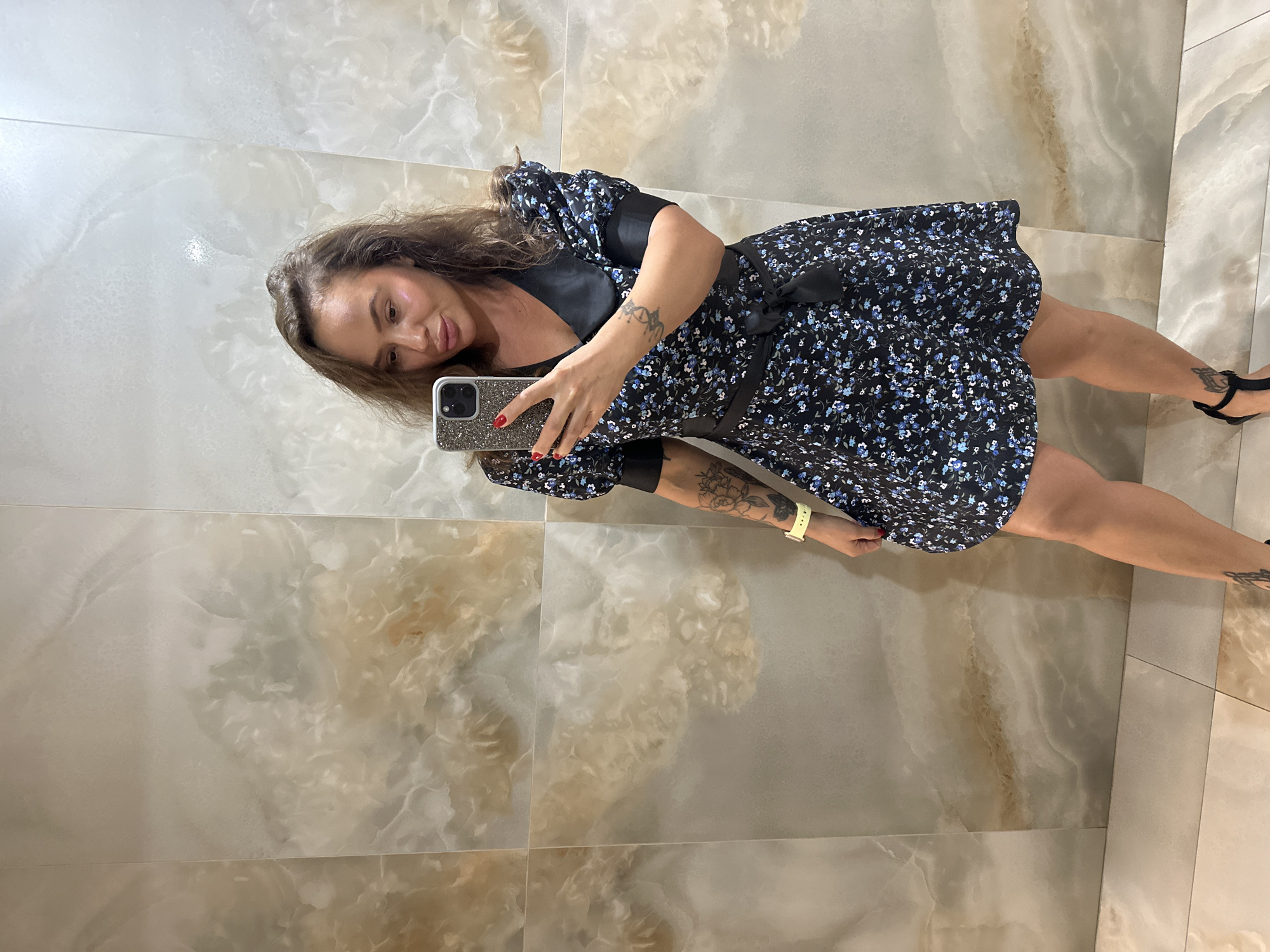 A woman takes a mirror selfie in what appears to be a restroom with marble-like tiled walls. She is wearing a short, black, floral-print dress with a black sash tied at the waist and a black collar. Her hair is medium-length and brown, and she is wearing red nail polish. She has tattoos on her left arm and calf. On her left wrist, she wears a yellow watch. She is holding a smartphone with a sparkly case. The background consists of large, beige and white patterned tiles.