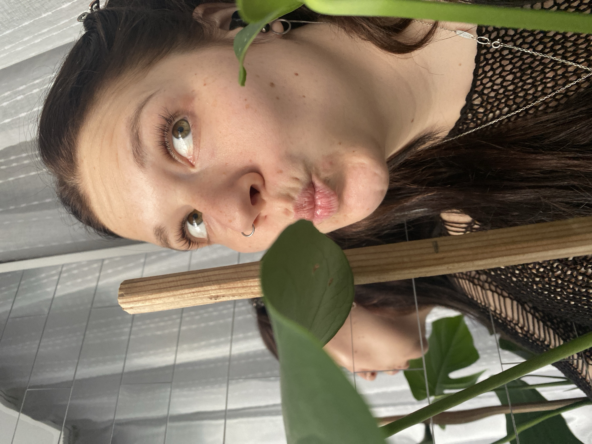 A young woman with fair skin, dark brown hair, and hazel or light green eyes is captured in a close-up, rotated portrait. Her head is positioned to the left, and she looks upwards with a slightly pouty or puckered lip expression, conveying a playful mood. Freckles are visible across her nose and cheeks. She wears a silver nose ring on her left nostril and delicate silver jewelry, including an ear cuff or small earring on her left ear and a thin chain necklace around her neck. Tiny, almost illegible text, possibly '925' indicating sterling silver, is visible on the necklace clasp.
She is wearing a black, open-knit or fishnet top. Several dark hair clips secure her hair on the top of her head. In the foreground, a large green leaf, likely from a Monstera plant, partially obscures the right side of her face. A smooth, light wooden stick or dowel is also visible horizontally in front of her lower face.
The background consists of a highly reflective surface, possibly a glossy tiled wall or a mirror, which shows fragmented reflections of the woman's face and the green plant. The overall lighting suggests an indoor setting with natural light illuminating her face. The image is aesthetically composed with the plant and wooden element creating a natural frame. The setting suggests an indoor environment, possibly a home, in Chaikovsky, Russia.