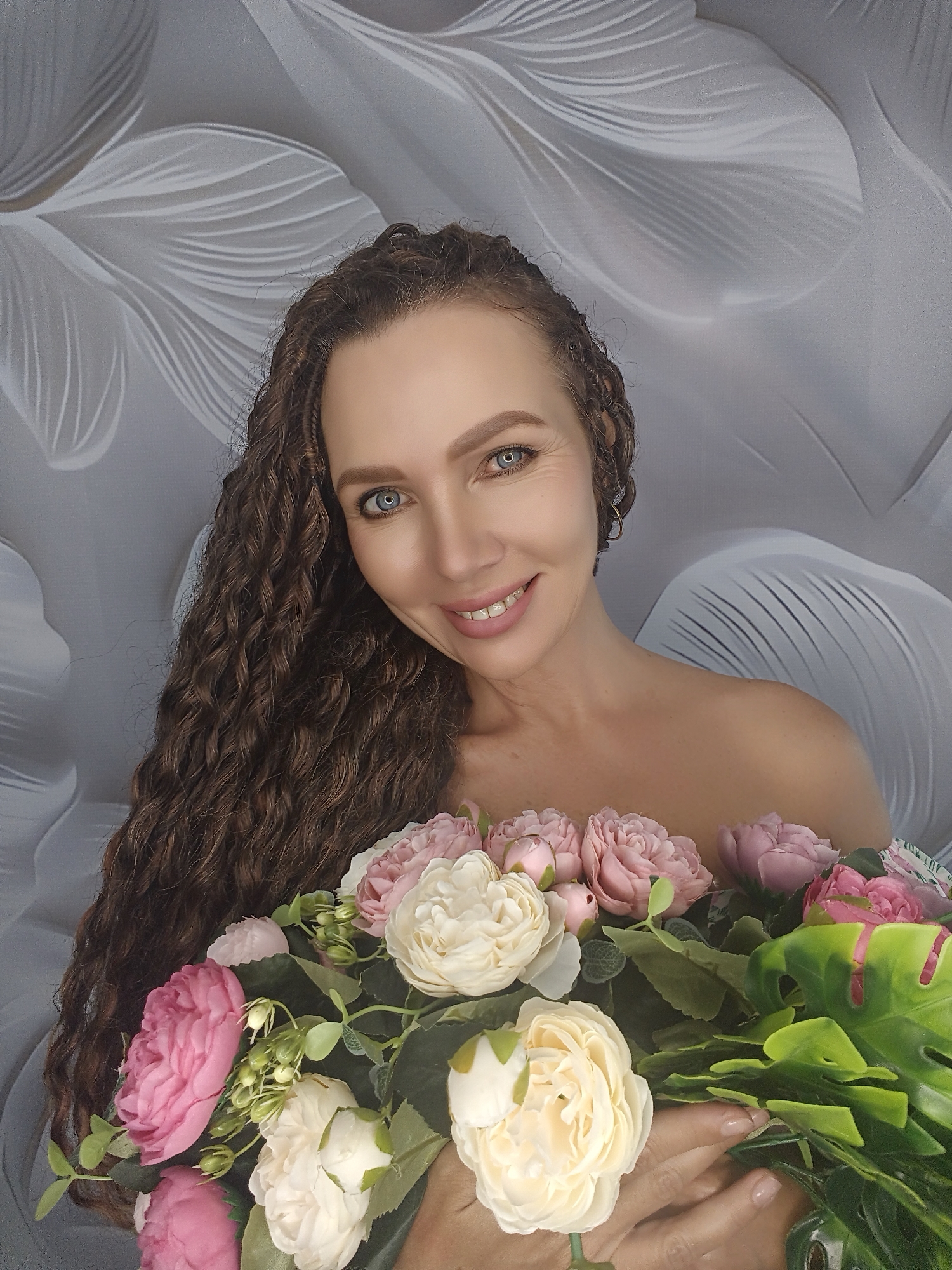 A woman with long, dark curly hair is holding a bouquet of pink and white flowers. She has bright blue eyes and a smile. She is wearing a light-colored top that exposes her shoulders. The background is a grey wall with a white floral pattern. The image is a portrait and is likely taken indoors.