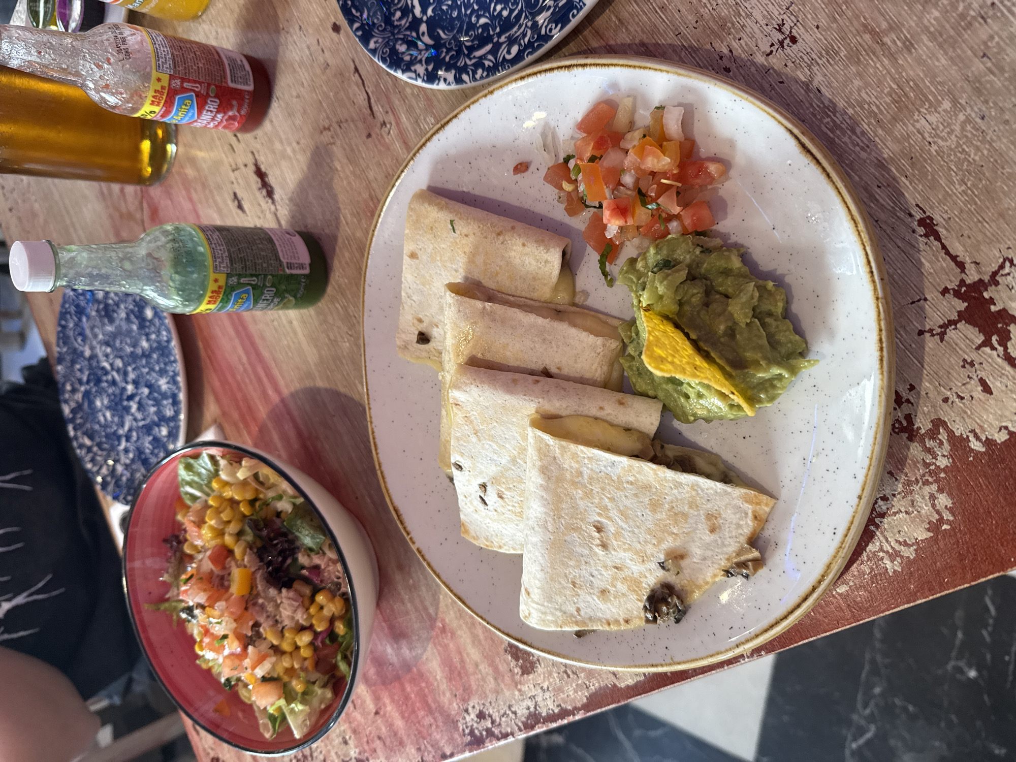 The image depicts a restaurant table in Madrid, Spain, laden with food. A white plate with a tan rim holds three quesadillas, guacamole topped with a tortilla chip, and pico de gallo. Adjacent to the plate is a bowl of salad containing lettuce, corn, tomatoes, and tuna. Three bottles of hot sauce are positioned to the left of the plate. A blue and white patterned plate is visible in the background. The table has a distressed wood-like surface, with areas of red and brown. A glimpse of a person's dark-colored shirt is visible on the left side of the image. Part of the floor is visible in the lower right corner, showing a pattern of black and white tiles.