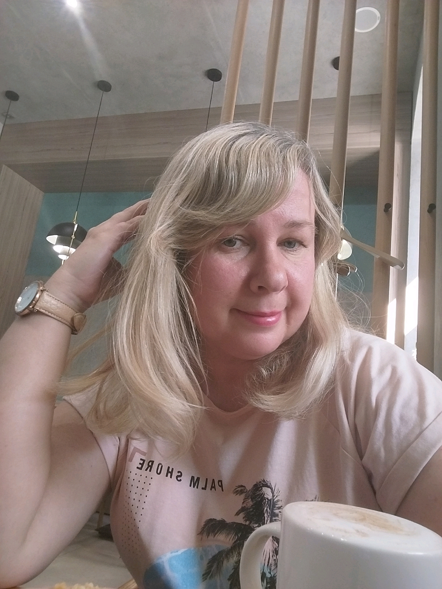 A woman with blonde hair is posing for a selfie in a brightly lit indoor space, possibly a cafe or a restaurant. She is wearing a light pink t-shirt with a palm tree graphic and text that reads "PALM SHORE" in reverse. On her left wrist, she wears a light brown watch. Her right hand is casually placed on her head, with her fingers running through her hair. In the foreground, a white mug with a frothy beverage, likely coffee, is visible. The background shows vertical wooden slats and decorative pendant lights hanging from the ceiling, suggesting a modern interior design. The overall lighting is warm and natural, with sunlight casting shadows through the wooden slats. There is no indication of the city of Orenburg, Russia in the image.