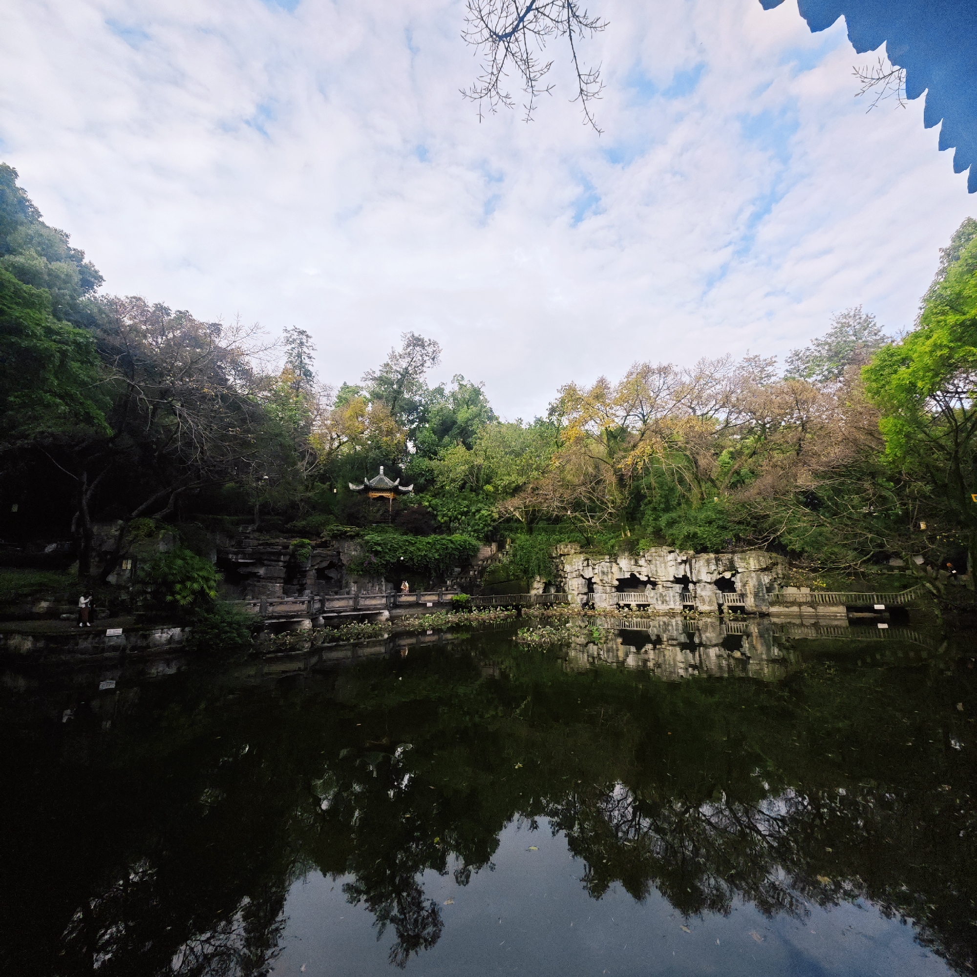 This media file captures a serene landscape, likely a classical Chinese garden or park, centered around a dark, reflective body of water. The scene is dominated by lush natural elements and traditional architecture.
In the background, a dense assortment of trees covers hilly terrain. Some trees display vibrant green foliage, while others exhibit autumnal hues of yellow and brown, or are bare, suggesting a transitional season like late autumn or early spring. Nestled among these trees on a higher elevation is a traditional Chinese pavilion with an ornate, dark-tiled roof, featuring pointed eaves.
Along the water's edge, intricate stone work forms a retaining wall, with sections featuring detailed rock formations and what appear to be carved grottoes or archways, especially prominent on the right side. Several narrow bridges and paved pathways, constructed from stone and wood, provide access along the perimeter of the water.
The water itself is dark and remarkably still, offering clear, albeit slightly blurred, reflections of the sky, the surrounding trees, and the architectural structures. Patches of aquatic plants, possibly lily pads, can be seen floating on the surface.
The sky overhead is bright, filled with a mix of soft white and grey clouds, with occasional glimpses of pale blue. The lighting is diffused, suggesting an overcast day or a time when the sun is not directly overhead, contributing to the tranquil atmosphere.
A few small figures, appearing to be visitors, are visible on the pathways by the water, adding a sense of scale and life to the expansive scene. One small, light-colored sign is present near the water, but its text is illegible. The traditional architectural style and natural setting are consistent with locations found in Yuzhong District, China.