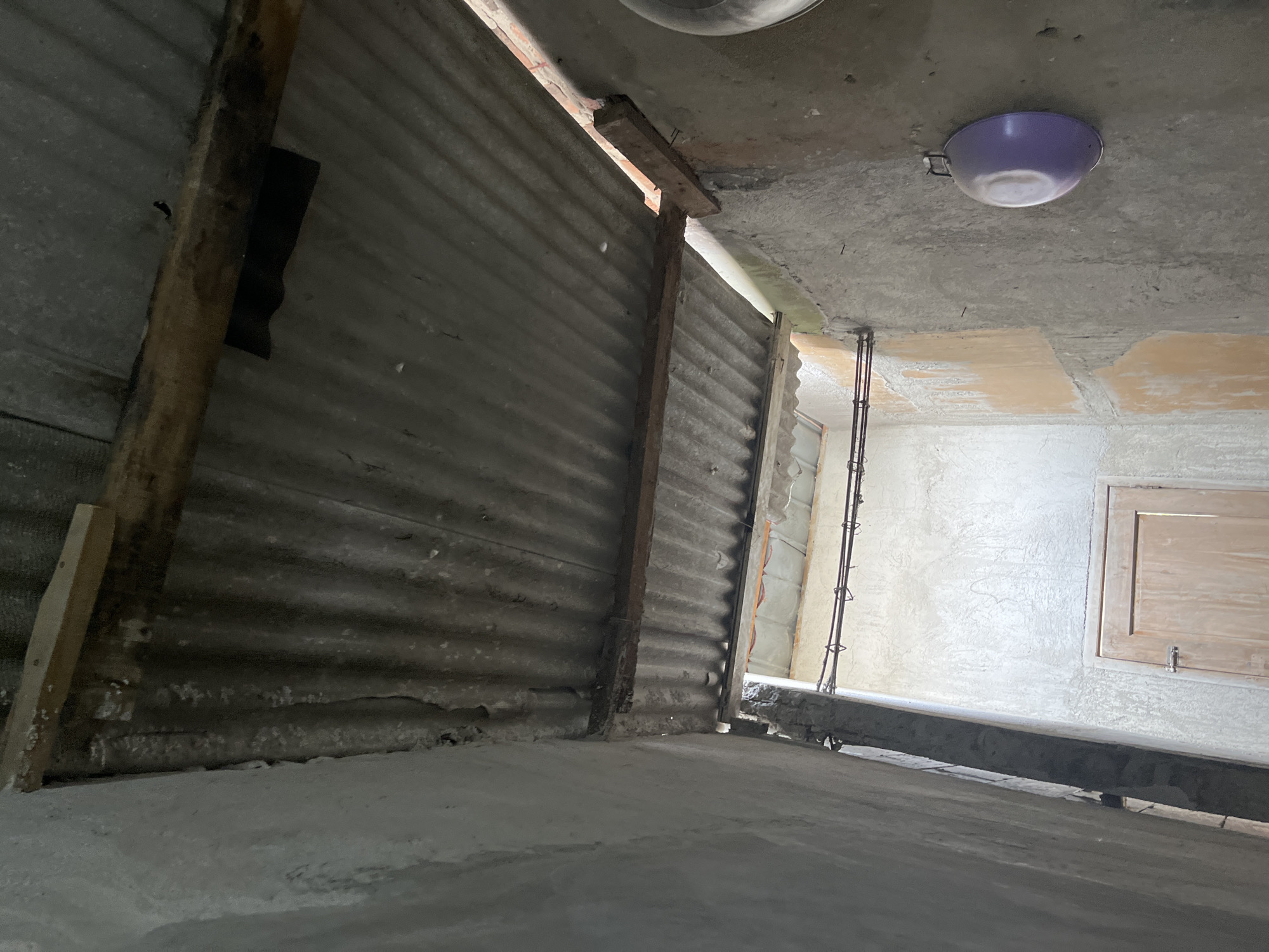 The image shows an interior view looking upwards, capturing a section of a rustic, unfinished structure. The ceiling appears to be made of corrugated metal sheets supported by wooden beams. To the left, a rough concrete wall is visible, curving slightly. On the right, another concrete wall is present, with a light purple basin hanging on it. Further into the scene, there is a wooden door with a metal latch and a visible hinge. The overall environment suggests a utilitarian space, possibly a shed or an outdoor kitchen area, in Laban, Indonesia. The lighting is dim, with some natural light filtering in from above. There are no people or animals present in the image.