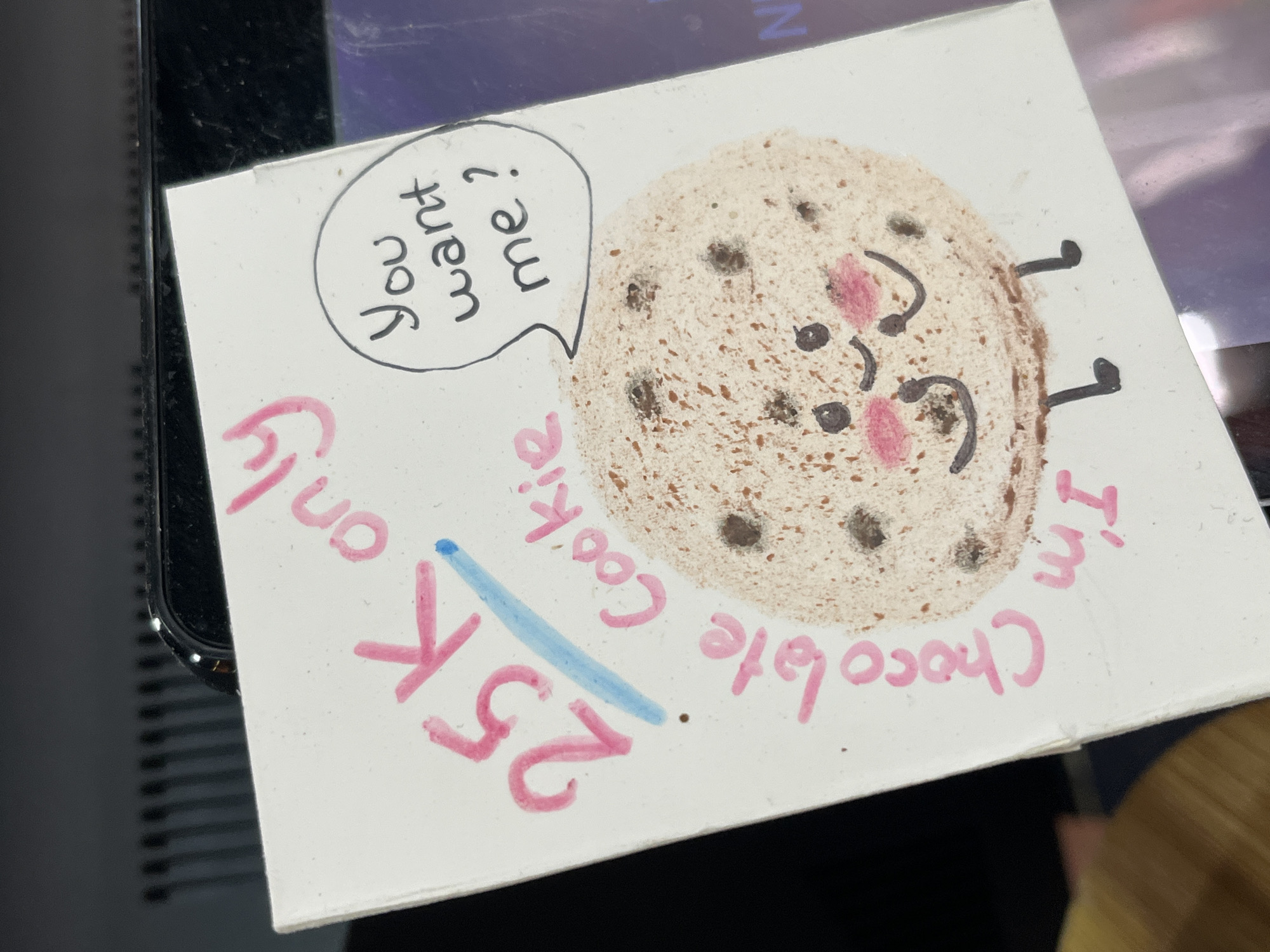 A small, square piece of white paper sits askew on a dark surface, likely a table or desk. The paper features a hand-drawn image of a chocolate chip cookie with a smiling face and two legs. A speech bubble above the cookie contains the text "You want me?" In pink marker, the text "I'm Chocolate Cookie only 25K" is written around the cookie. The blue underline indicates the price.
There is no information from the image to identify the location as Đà Nẵng, Vietnam.