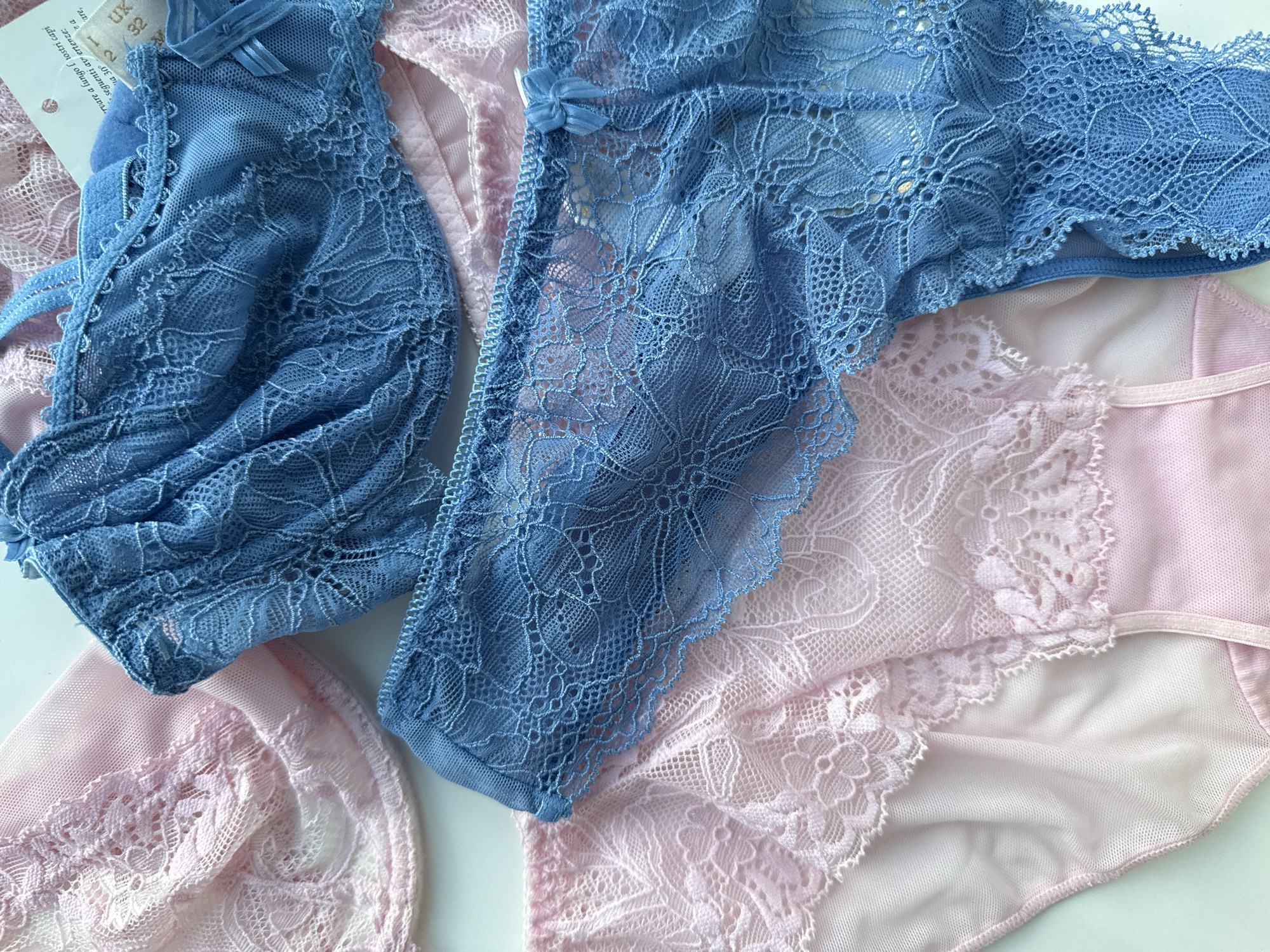 A top-down, close-up shot displays delicate lace lingerie in soft pink and vibrant blue. The pink pieces, likely a bra and panties, feature intricate floral lace trim overlaying a sheer fabric and opaque material. The blue pieces, a bra and thong, are crafted from a bolder floral lace with ruffled edges and a small satin bow on the thong. The lingerie is arranged on a clean, white surface, suggesting a studio or retail setting. No people or specific locations are visible.