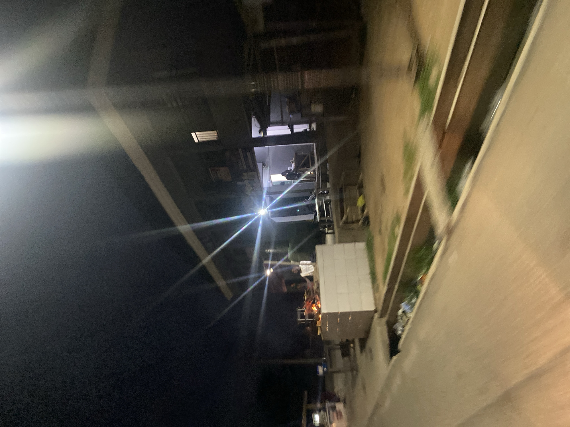This image depicts an outdoor scene at night, likely in a part of Jalingo, Nigeria. The foreground is dominated by a blurred street or pathway, suggesting movement, possibly from a vehicle. To the right, a drainage channel is visible, containing some debris and grass. Further back, a building with a light on inside is present, with what appears to be a shop or establishment. In front of this building, a raised, white structure, possibly a counter or display, is prominent. On top of this structure, a fire is burning, with several pieces of wood or materials being consumed. A person, dressed in light-colored clothing, stands behind the fire. Another person can be seen inside the building. The overall atmosphere is dark, with artificial lighting from the building and the fire creating strong highlights and lens flares. The environment appears to be an urban or semi-urban setting, characterized by the building, paved surfaces, and visible infrastructure.