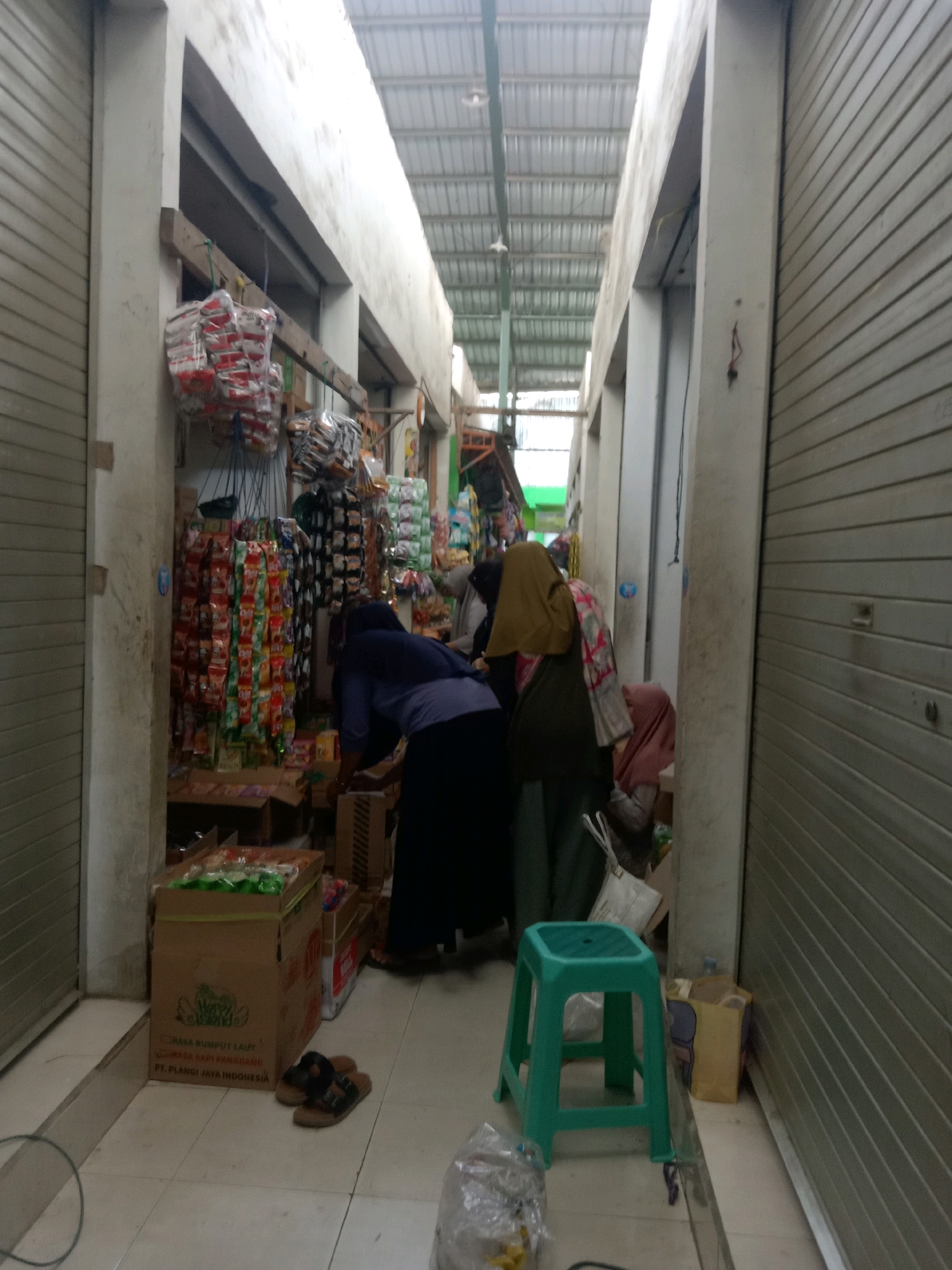 The image depicts a narrow indoor market aisle in Sugihwaras, Indonesia. The aisle is flanked by roll-up metal doors on both sides, suggesting a shopfront. The ceiling is made of corrugated metal. On the left, stacked shelves display numerous small, colorful packets of what appear to be snacks or convenience food items. A few individuals, presumably shoppers or vendors, are present. One person in the foreground is bent over, interacting with items on a lower shelf. Another person, wearing a headscarf and a dark garment, is partially visible. In the middle of the aisle, a bright green plastic stool sits on the tiled floor. Several cardboard boxes are stacked around the market stalls, some containing merchandise. On the floor, a pair of sandals is visible near a large cardboard box. The overall scene conveys a bustling, albeit somewhat cluttered, retail environment.
