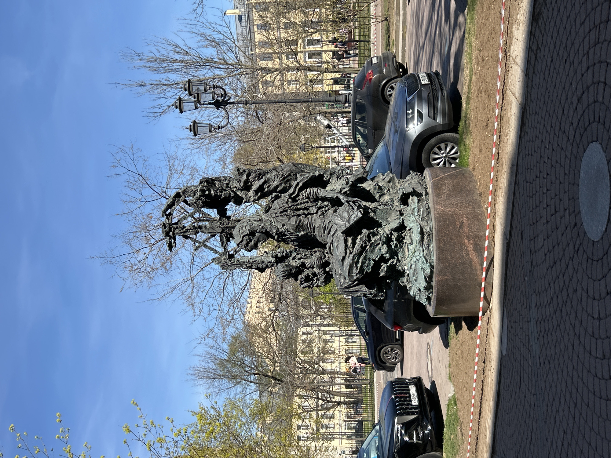 The image depicts a bronze sculpture on a stone base, possibly a monument or public art installation, in Saint Petersburg, Russia. Several cars are parked along the side of the sculpture, suggesting it is located in a street or parking area. Behind the cars and sculpture is a fenced-off area with trees and a building. The sky is clear and blue. A street lamp is visible to the left of the sculpture. The sidewalk and street are visible, suggesting an urban environment. A white and red striped tape is near the edge of the sidewalk.