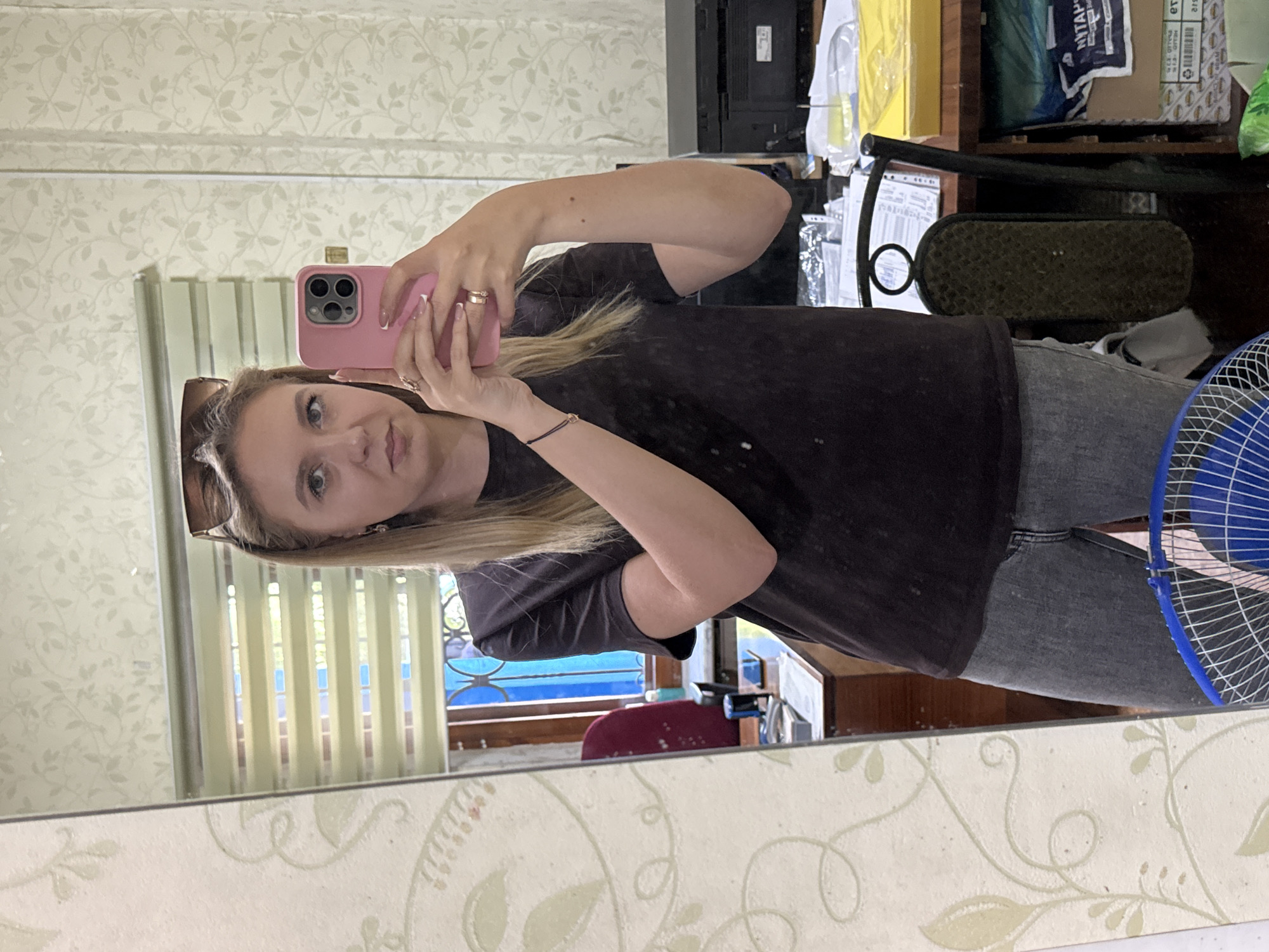 In a room with patterned wallpaper, a young woman with blonde hair and sunglasses perched on her head is taking a selfie in a mirror. She is wearing a dark t-shirt and grey jeans, and is holding a pink smartphone in her right hand. The reflection in the mirror shows a desk with papers, a printer, and a chair. The room appears to be an office or study area.