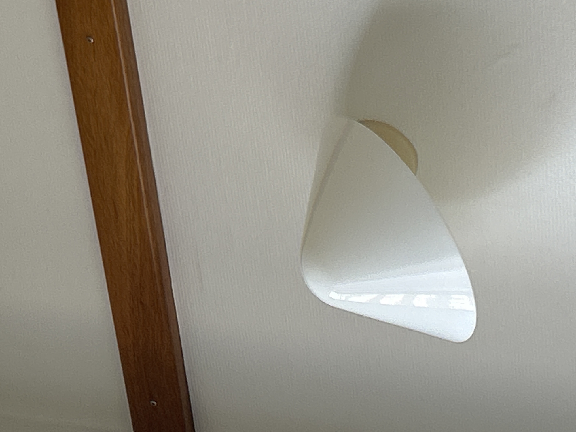 The image shows a close-up of a wall with a wooden trim running horizontally across the top. Below the trim is a white wall with a textured surface. Mounted on the wall is a white, curved light fixture. The light fixture has a smooth, reflective surface that shows reflections of what appears to be windows or lights. A portion of a circular, yellowish object is visible within the opening of the light fixture. The scene does not contain any people or specific identifiable landmarks that would place it within the city of Gummersbach, Germany.
