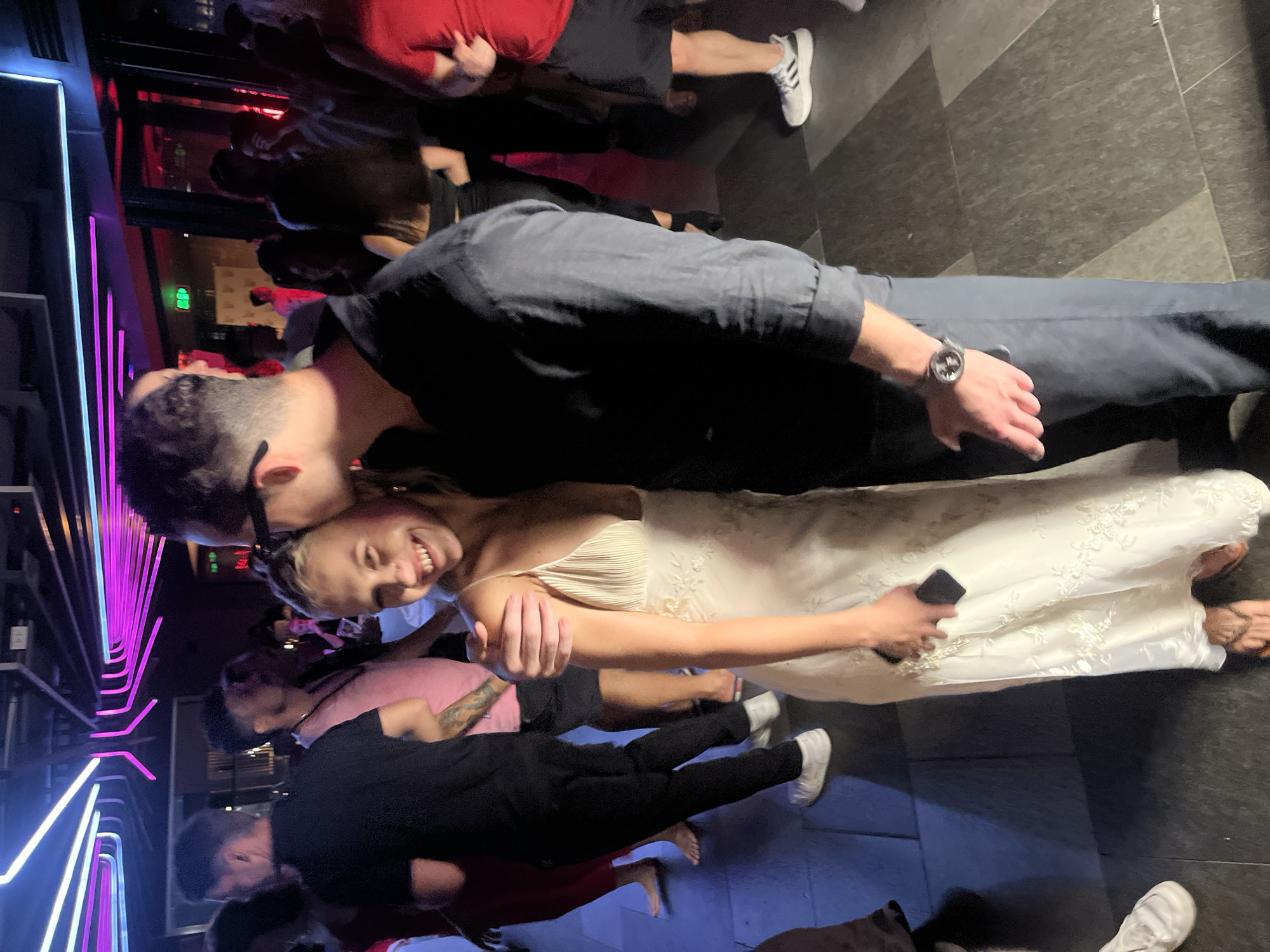 The image captures a candid moment of a couple at what appears to be a wedding reception or a celebratory event. The couple is the primary focus, with the groom leaning in to kiss the bride. The bride is wearing a white, intricately detailed wedding dress with pleats on the bodice and floral embroidery on the skirt. She has a joyful expression, smiling broadly. The groom is dressed in a dark shirt and pants and has glasses.
Several other people are visible in the background, some dancing or socializing, suggesting a lively atmosphere. The setting seems to be an indoor venue, possibly a club or a banquet hall, with distinctive neon lighting in shades of pink and purple along the left side of the image. The floor is tiled with a dark, patterned material.
The image appears to have been taken at night, given the artificial lighting and the indoor setting. The overall mood is celebratory and romantic, with the focus on the couple's affection. No visible text is present in the image. The location context of Da Nang, Vietnam, is provided but not directly evident from visual cues within the image itself.
