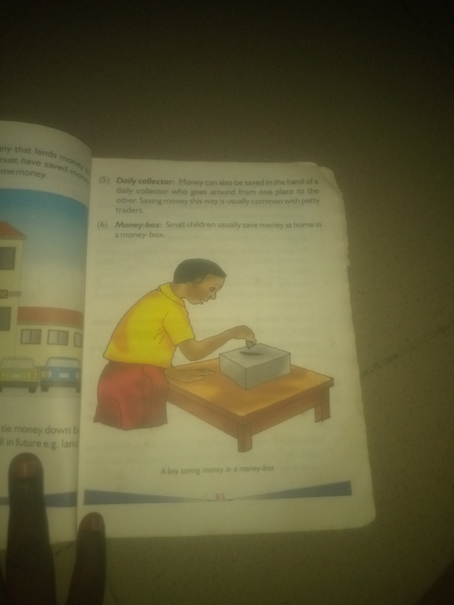 The image shows an open book with text and an illustration. The text on the left side discusses lending money and saving. The right side of the book contains two numbered points. Point (5) describes a "Daily collector" who saves money, and point (6) discusses "Money-box" and how small children save money at home. Below the text is an illustration of a young boy in a yellow shirt and red shorts, looking down at a grey money box on a wooden table. He is in the process of putting something into the slot on the money box. The background of the illustration shows a building and two cars. The text below the illustration reads "A boy saving money in a money-box." The page number "83" is visible at the bottom right. The image is taken from a high angle, and parts of two fingers are visible in the foreground. The scene depicted is not specific to Jalingo, Nigeria, or any particular city.