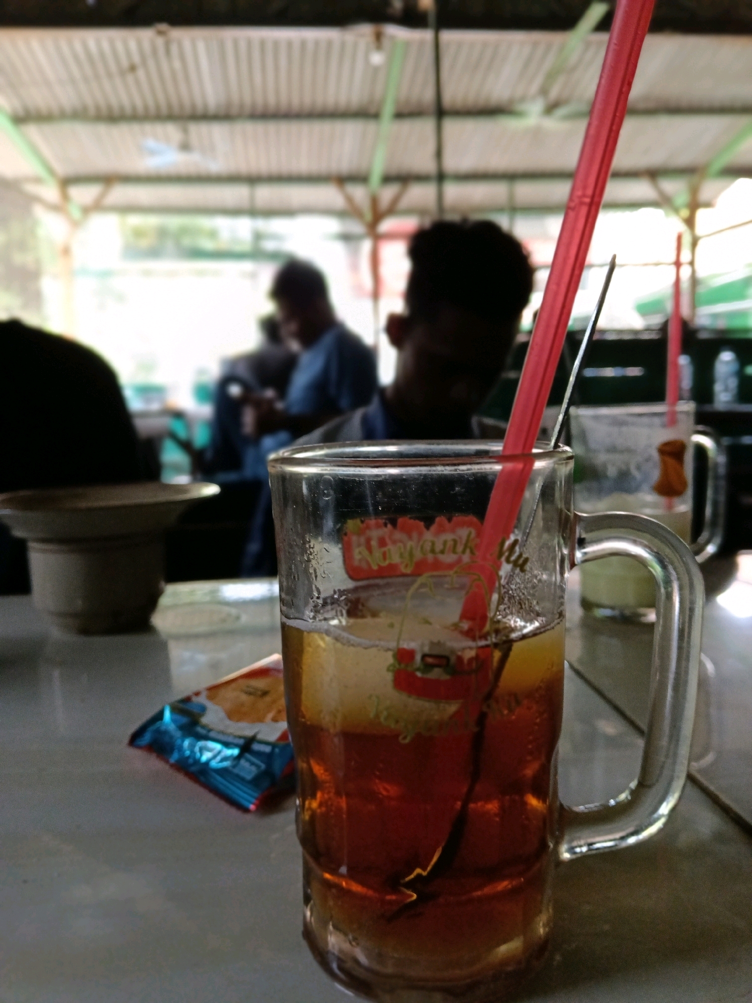 A close-up, shallow depth-of-field shot focuses on a glass of iced tea with a red straw and a metal spoon submerged within it. The glass sits on a textured, light-colored surface, possibly a table. To the left of the glass, slightly out of focus, is a small, crinkled, blue and orange package, possibly containing snacks. Behind the glass, other people and objects are blurred, suggesting an outdoor or semi-outdoor seating area. The background reveals a corrugated metal roof supported by green beams, characteristic of a simple open-air structure, likely a warung or a casual eatery. The lighting appears natural, possibly from overhead. The overall scene suggests a relaxed, everyday moment, possibly in the city of Blora, Indonesia, given the cultural context of such casual dining establishments.
