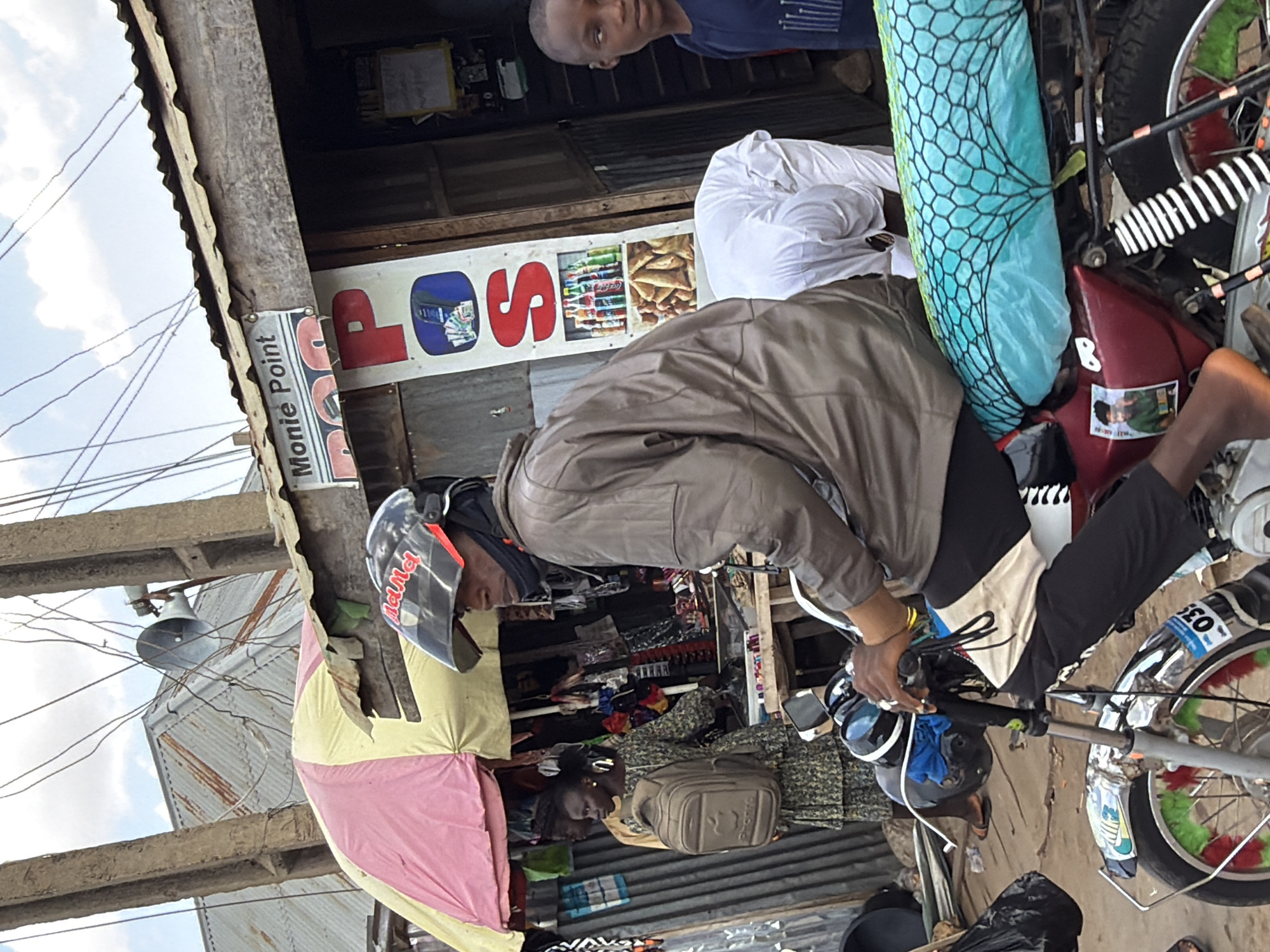 A street scene in Jos, Nigeria shows a man on a motorcycle in front of a small store. The store's sign reads "LOS" followed by an image of products. The motorcycle has a turquoise net strapped to the back and white and black accents on the rear fender. A man in a white shirt is standing behind the seated man, along with a younger man looking towards the camera in the doorway of the store. An umbrella is on the left side, partially obstructing the view.