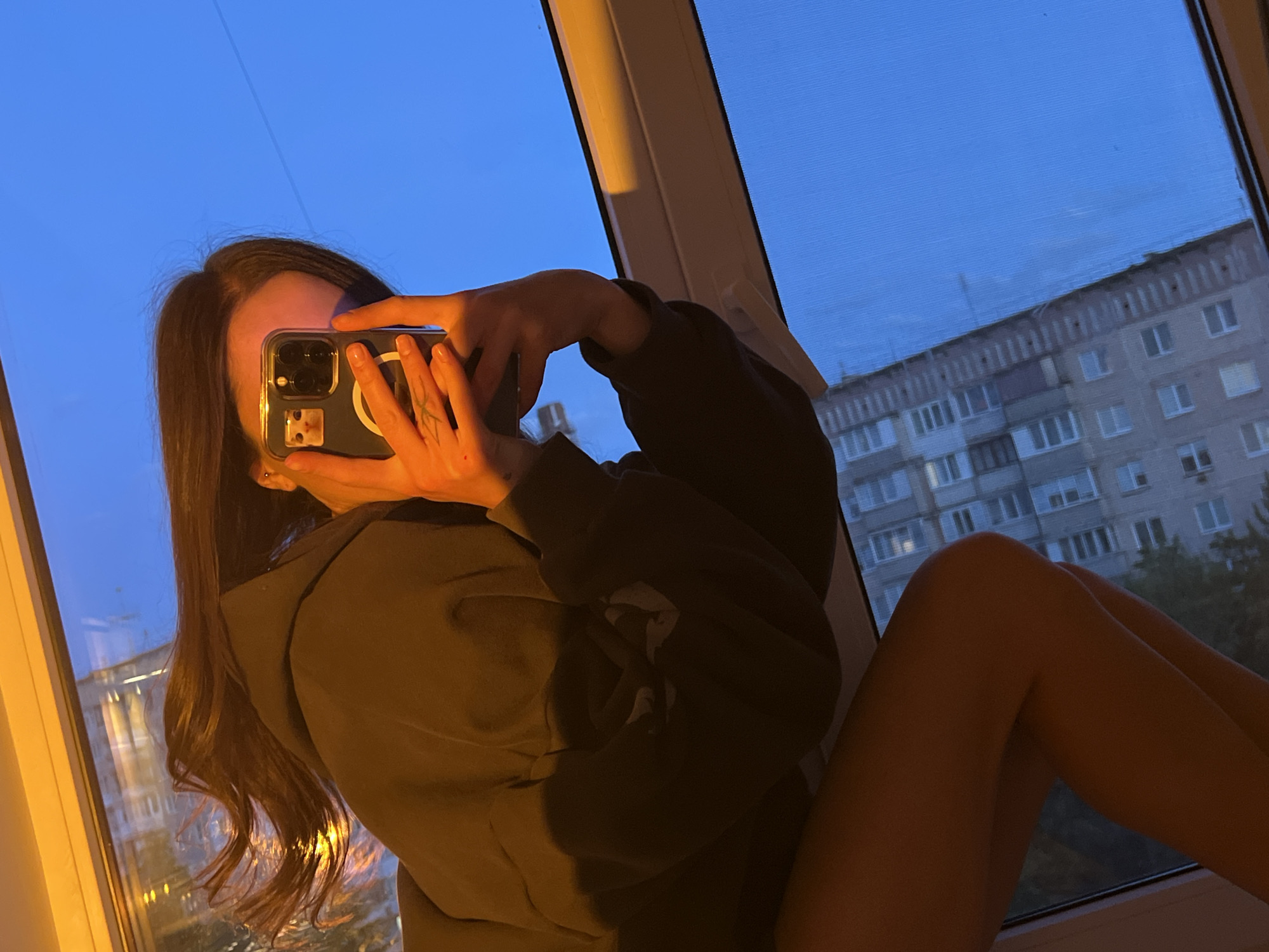 A young woman sits in a window, taking a picture with her phone. The window offers a view of a multi-story building across the street. It is dusk or twilight, with a blue sky visible. The woman has long, dark hair and wears a dark hoodie. Her legs are visible, suggesting she is sitting with her knees pulled up. The style of the building across the street suggests the location could be Ternopil, Ukraine.