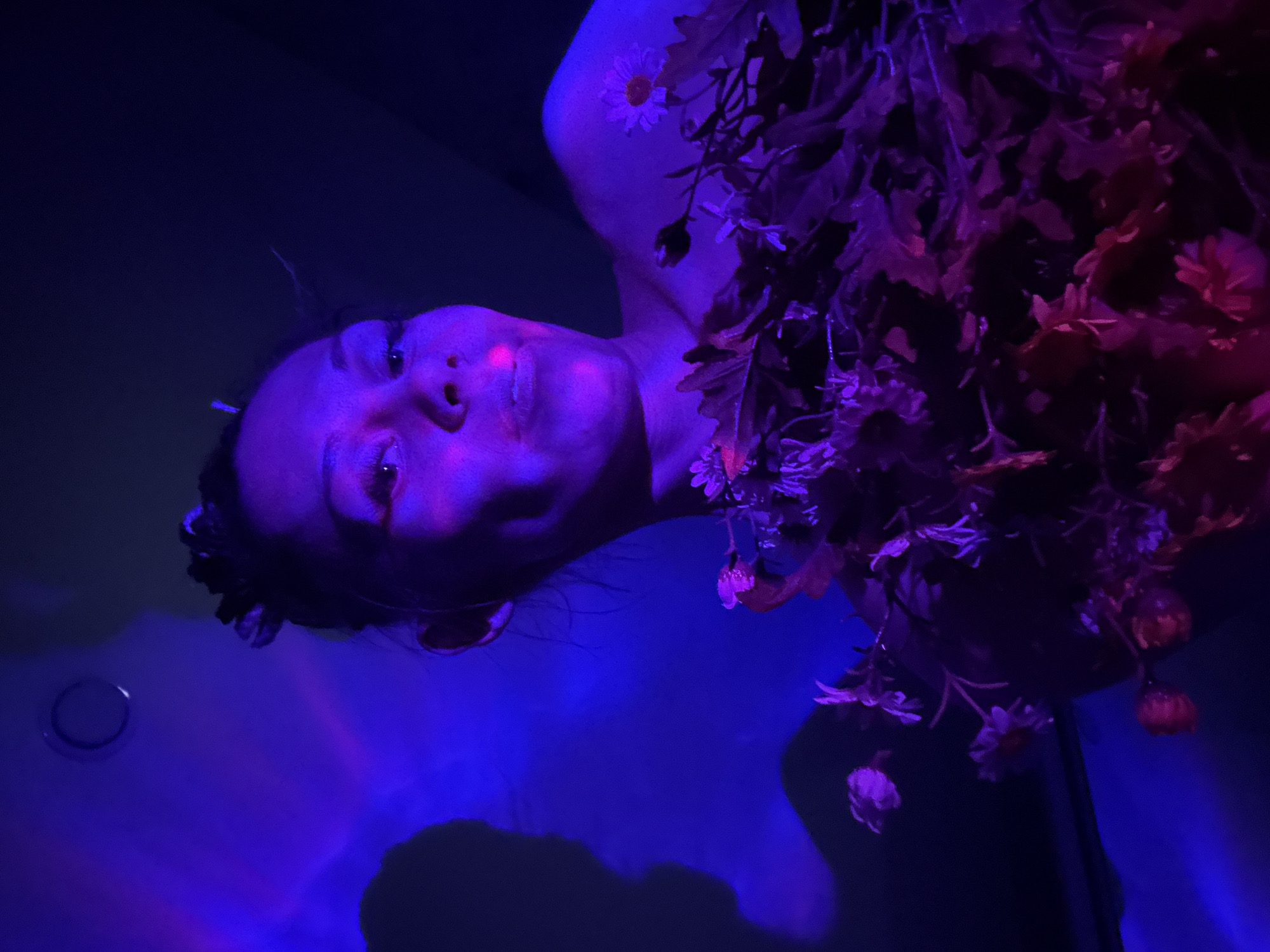 A person lies in a bathtub filled with water, photographed from above in a dimly lit room, likely in Rusanovo, Russia. The lighting casts a blue hue across the scene, with hints of purple and red around the person and the floral arrangements that adorn them. The person has a bare shoulder visible, with a single white daisy resting on it. A profusion of mixed artificial flowers and foliage surrounds the person, partially obscuring them. A circular object is visible on the side of the bathtub. The image is oriented so that the person's head is near the bottom left corner, and their body extends upwards and to the right. The person's face is visible, appearing relaxed. Their hair is dark and slightly disheveled. The lighting and floral decorations suggest an artistic or themed setting.