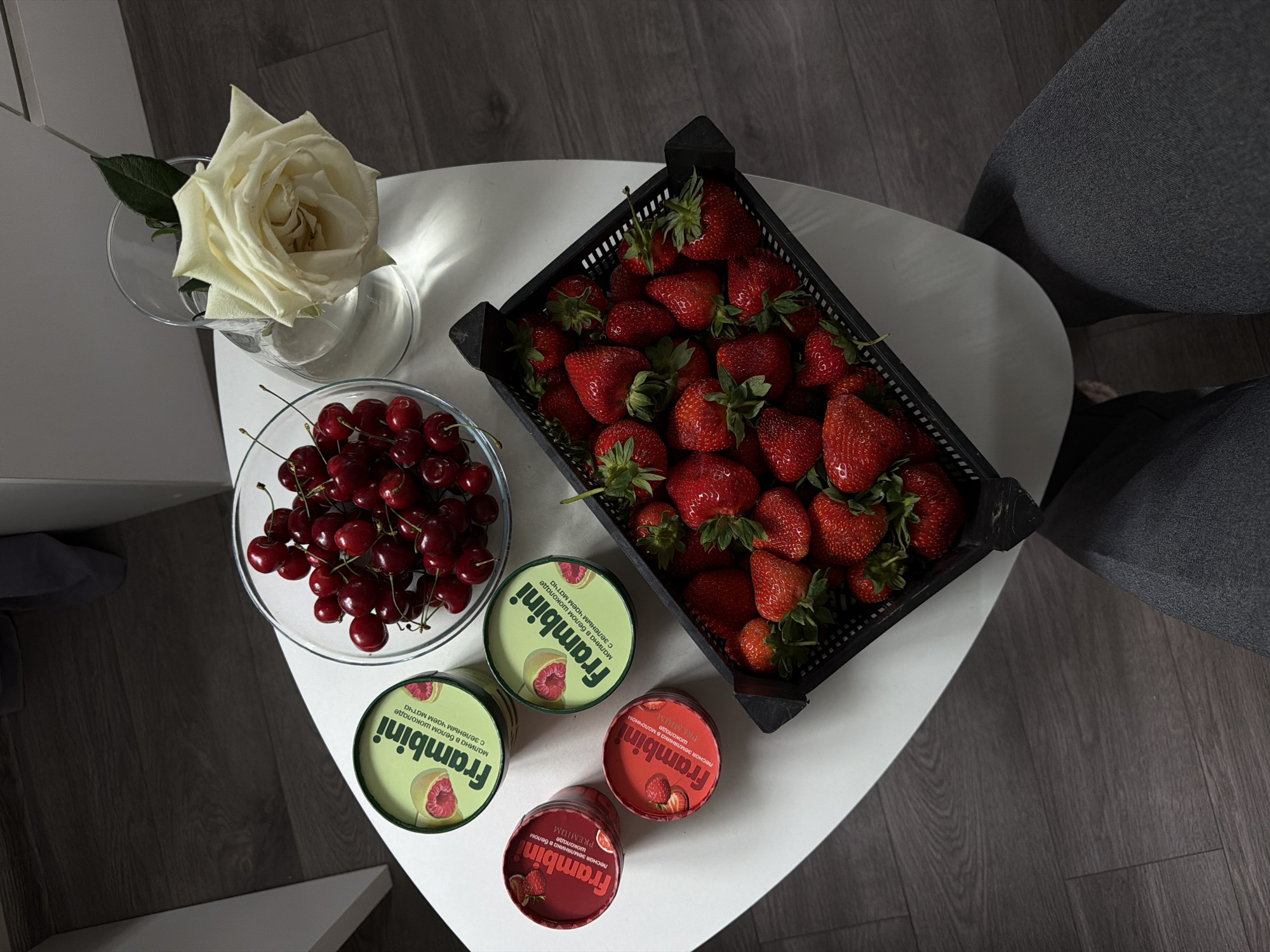 The image shows a still life arrangement on a white triangular table, possibly indoors. The table rests on a dark wooden floor, adjacent to a grey upholstered chair. On the table are: a clear glass vase holding a white rose, a clear bowl filled with dark red cherries, a black plastic crate overflowing with ripe strawberries, and four small containers of a product labeled "Frambini" in Russian, indicating chocolate or jam flavored with various berries like raspberry and strawberry. The scene is lit from above, casting shadows. No people are clearly visible, though part of a person's legs and dark-colored clothes are present near the chair. The image does not contain information to indicate the city of Kazan.