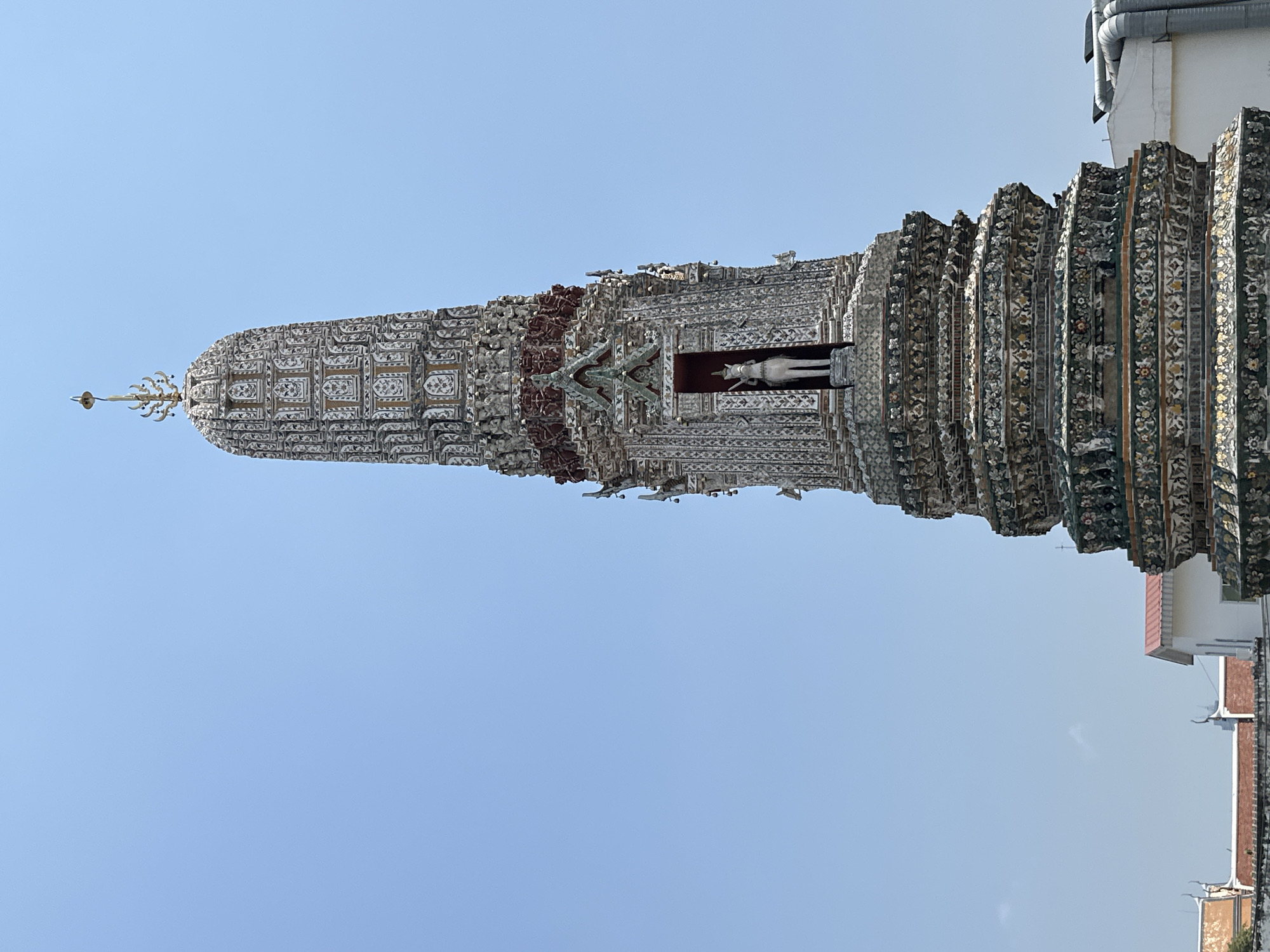 The image shows a vertical, upward view of the iconic Wat Arun Ratchawararam Ratchawaramahawihan, or Temple of Dawn, in Bangkok, Thailand. The central prang (tower) is the main subject, intricately decorated with colorful glazed ceramics and seashells. A statue of a deity is visible in a niche of the prang. The structure is set against a clear, light blue sky. The lower right corner shows part of adjacent buildings. No people are visible in the shot.
