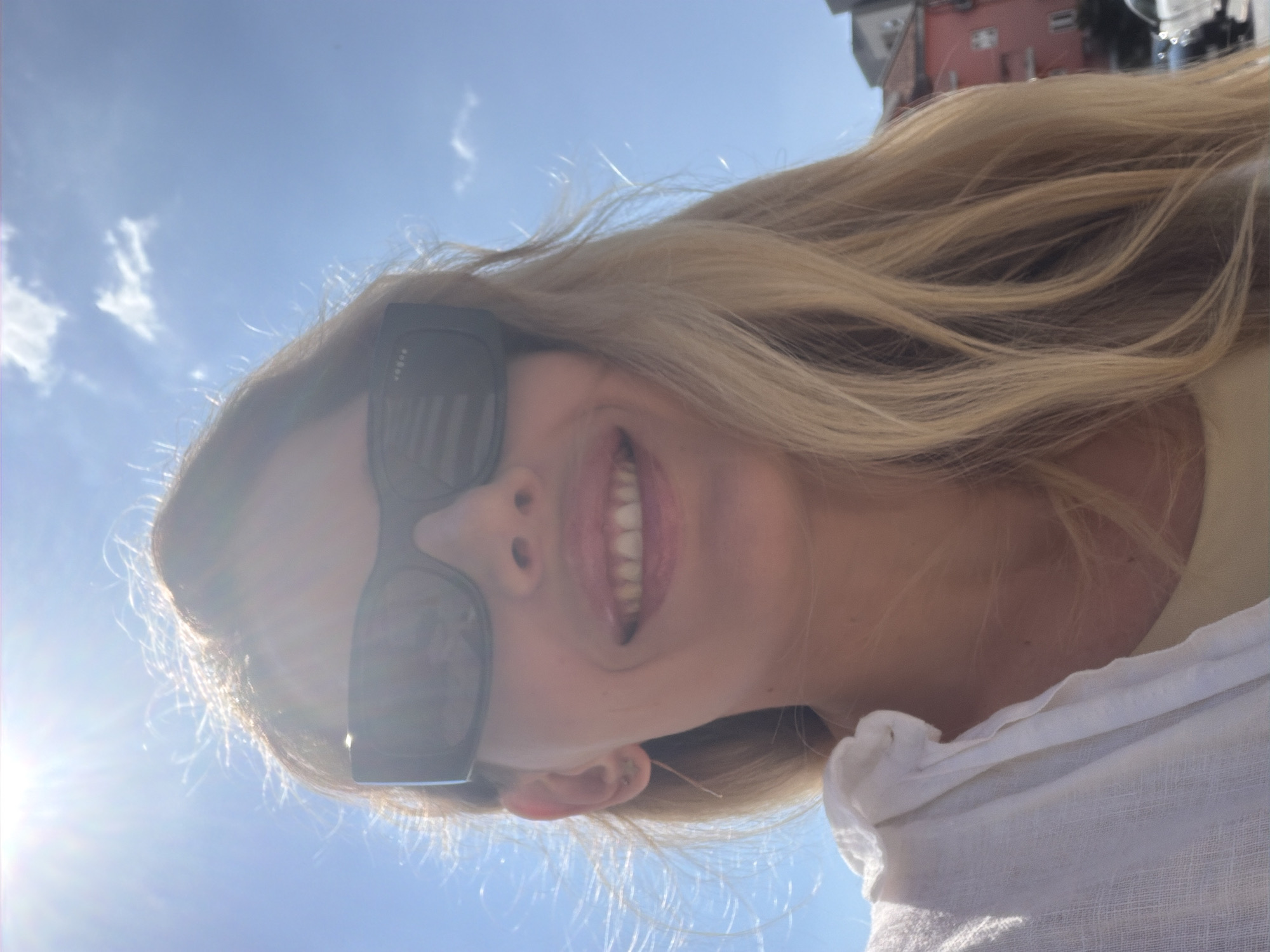 The media file is a portrait of a young Caucasian woman with long blonde hair, taken outdoors during the daytime. She is wearing dark sunglasses and a light-colored top. Her mouth is open in a smile, revealing her teeth, and her eyes are hidden behind her sunglasses. The background is a bright blue sky with some scattered white clouds, suggesting sunny weather. The sun's glare is visible on the left side of the image, indicating it's likely late morning or early afternoon. There are some out-of-focus buildings in the upper right portion of the image, with red and beige colors, hinting at an urban or coastal setting. The woman's hair is flowing, possibly due to a gentle breeze. The overall mood appears cheerful and relaxed. No visible text is present in the image. The style of the buildings and the sky conditions are consistent with a location like Budva, Montenegro.