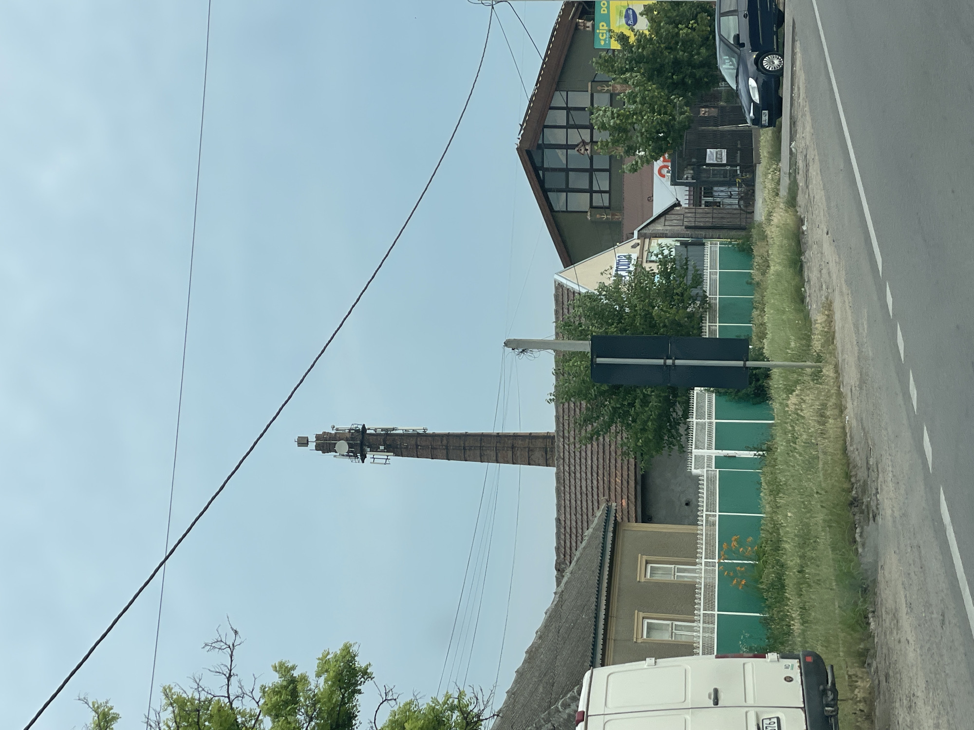 In Congaz, Moldova, a scene unfolds beneath a cloudy sky, featuring a towering brick chimney converted into a telecommunications tower. The tower, adorned with antennas and various equipment, stands prominently against the overcast heavens. At the base of the chimney, residential buildings with tiled roofs are visible, some featuring traditional architectural elements. A white van is parked on the left side of the image, and a dark blue car is positioned on the right, near the edge of a paved road with white markings. A green fence separates the buildings from the roadside, with overgrown grass and vegetation growing in front of it. The overall impression is one of a rural or semi-urban environment where old industrial structures have been repurposed for modern technological use.