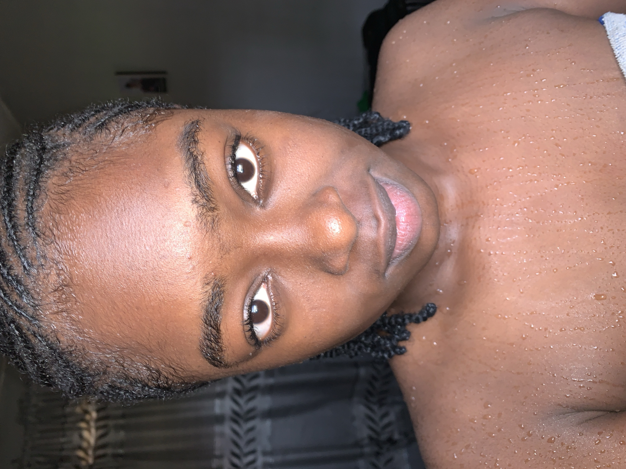 This close-up portrait features a young individual with dark skin, positioned with their head tilted and looking directly at the camera. Their hair is styled in tightly braided cornrows that frame the face and extend back. Numerous small water droplets are visible on the skin across the face, neck, and upper torso, suggesting the person has recently showered or been in contact with water.
The subject's eyes are open, revealing dark brown irises and well-defined eyelashes. Their lips are slightly parted, showing a hint of the inner lip. A natural sheen from the water and skin oils is apparent across their complexion. Part of a blue and white striped towel or fabric is visible on the right side of the image, near the subject's shoulder. The background is dark and out of focus, with a faint, blurry rectangular object visible in the upper left, possibly a picture or frame on a wall. The person maintains a neutral expression. There is no visible text in the image.