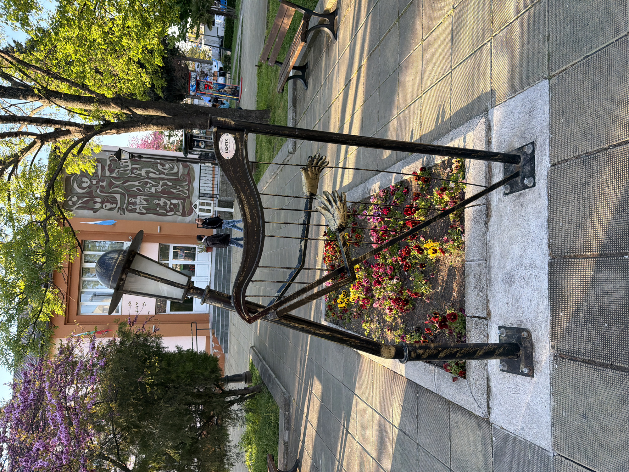 Here is a detailed description of the image:
The image shows an outdoor scene featuring a decorative metal structure with two sculpted hands reaching out from the center, situated within a flowerbed filled with pansies. The metal structure seems to be part of a street decoration or possibly a memorial in an urban setting. It includes an attached old-fashioned street lamp. In the background, there is a building with a mural that has abstract figures, trees, benches, and people walking on the sidewalk. The ground is covered with textured paving stones and sunlight is casting shadows across the scene. The location appears to be a public park or square in Unknown, Unknown.
