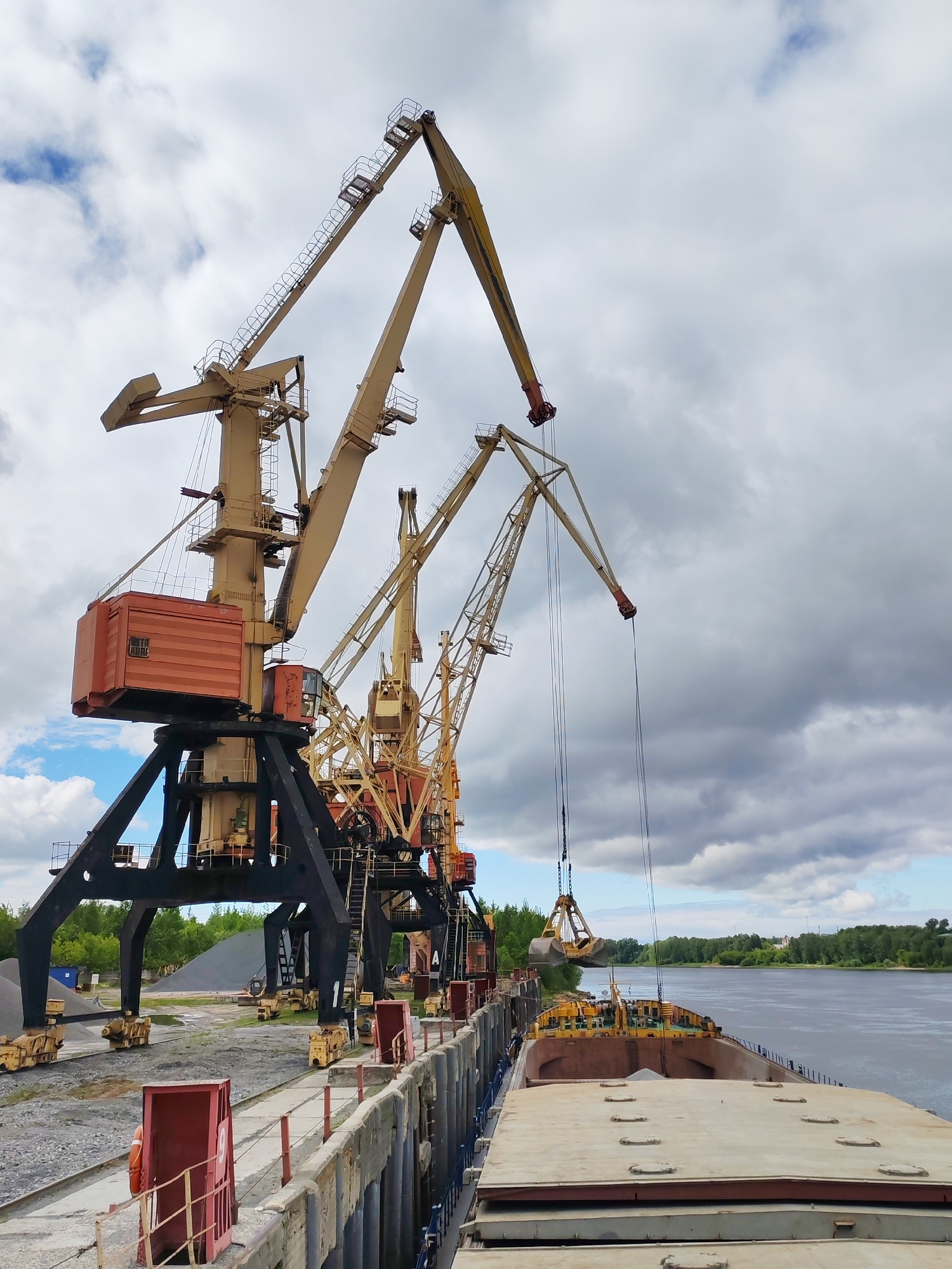 The image shows a scene at a port in Rybinsk, Russia. The sky is overcast with grey clouds. Several large, industrial cranes dominate the view, painted in beige and black, positioned along a concrete pier. The cranes are actively engaged in loading cargo onto a barge, with one crane's grab bucket suspended over the open hold of the barge, presumably filled with material. The barge is also yellow. The river is visible in the background. A strip of green trees lines the opposite bank of the river. On the pier, there are safety measures in place, with open structures that are red/orange with red bars that serve as safety gates. Behind the cranes, there are piles of gravel, and a faint treeline.